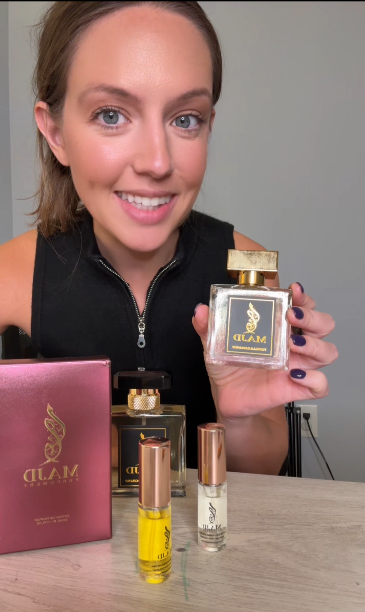 A woman with short brown hair and blue eyes smiling at the camera while holding a perfume bottle. Several other perfume bottles and a pink box are on the table in front of her.