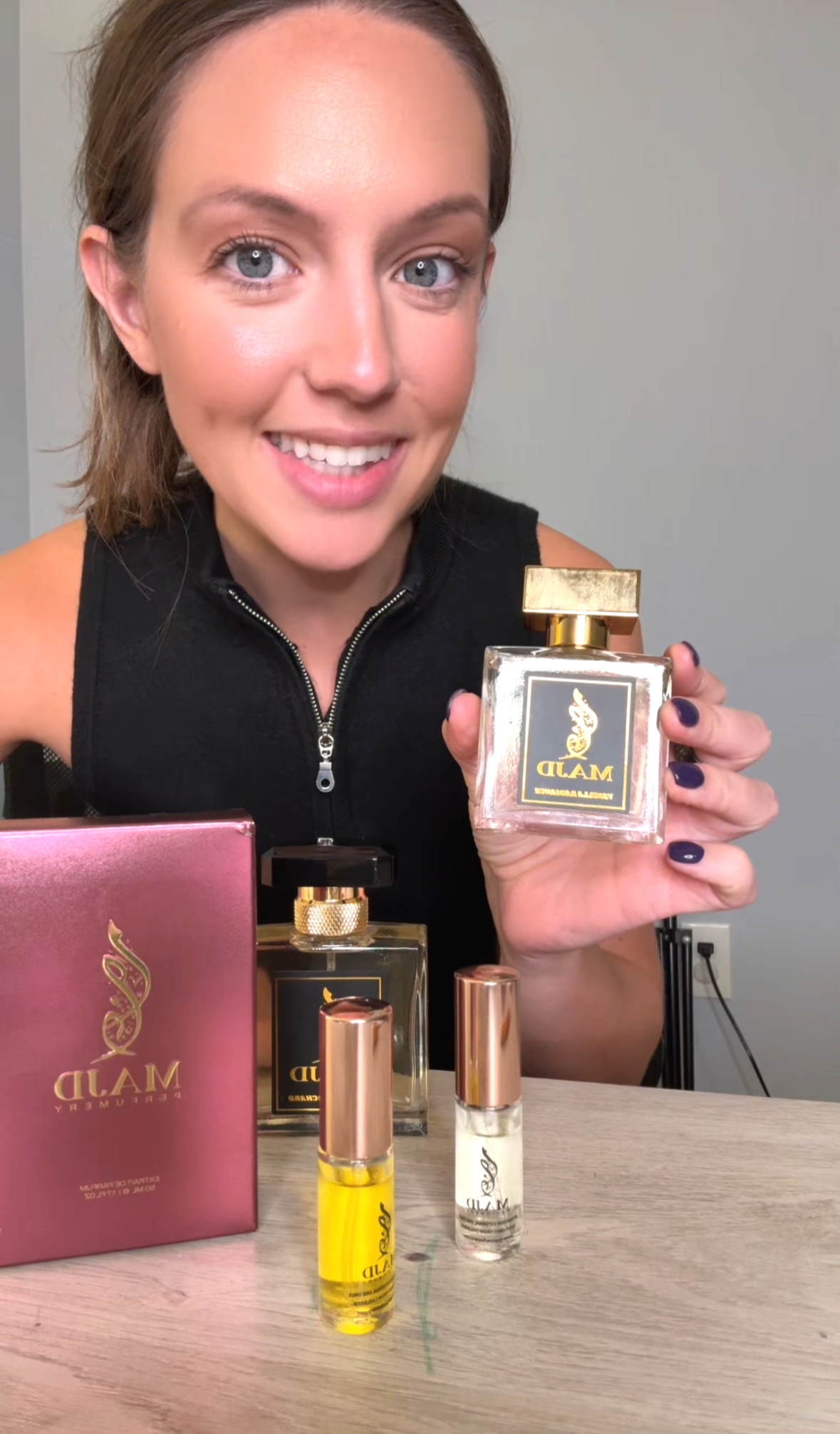 A woman with brown hair and blue eyes smiling at the camera while holding a perfume bottle. On the table in front of her are several perfume bottles and a pink box, all branded with the 'MALD' logo. The background is plain and unadorned.
