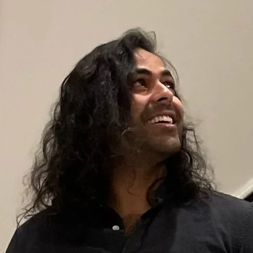 Man with long, curly black hair smiling and looking upward, wearing a dark shirt against a plain wall background.