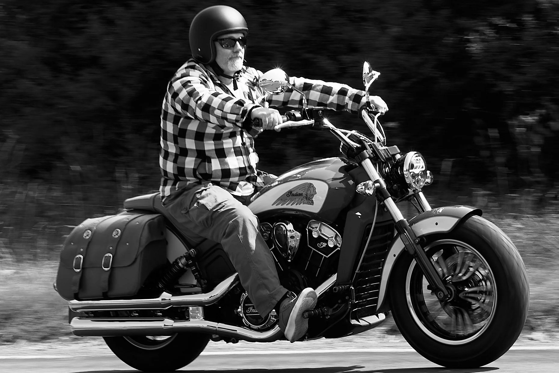A man wearing a helmet, sunglasses, and a checkered shirt riding an Indian motorcycle with saddlebags, moving along a road with a blurred background.
