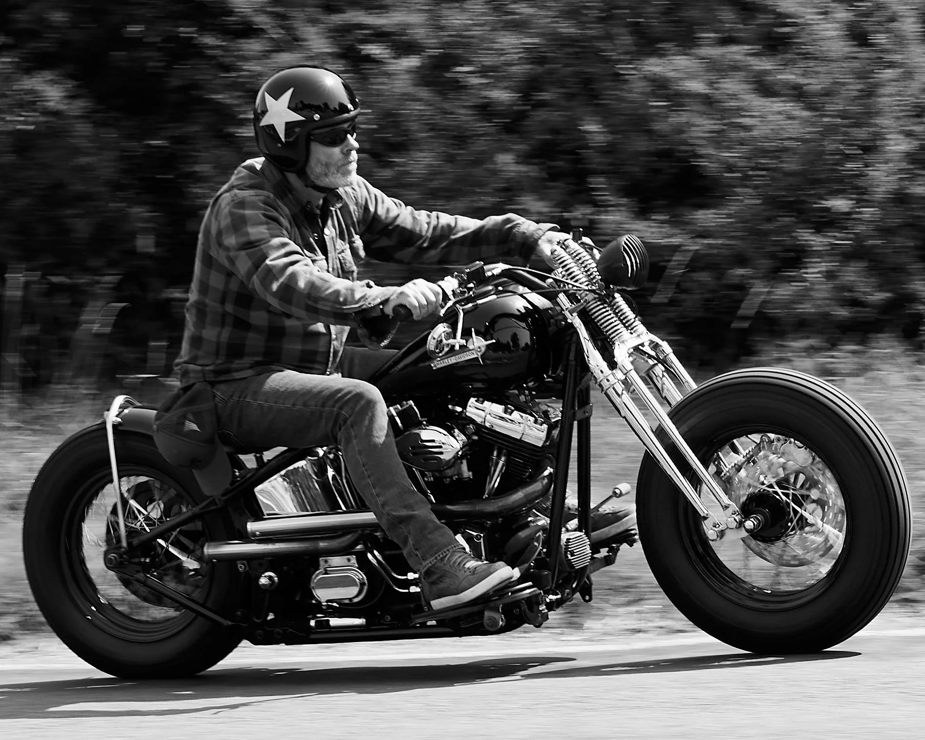 A man with a beard riding a motorcycle on a road. He is wearing a helmet with a star on it, sunglasses, a plaid shirt, jeans, and sneakers. The photo is in black and white.