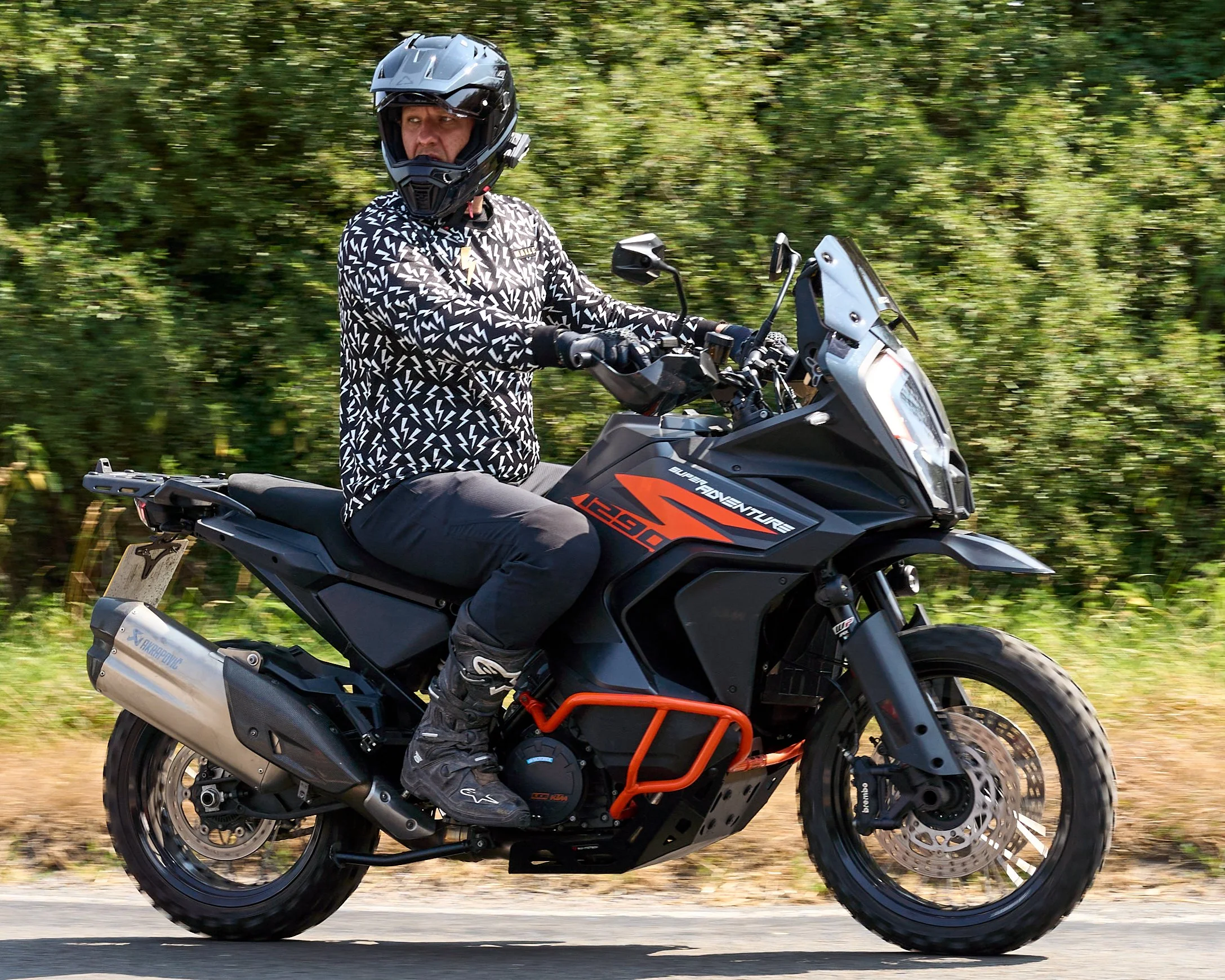 A person wearing a black full-face helmet, a black and white patterned jacket, and black gloves riding a black and orange adventure motorcycle on a paved road with green foliage in the background.