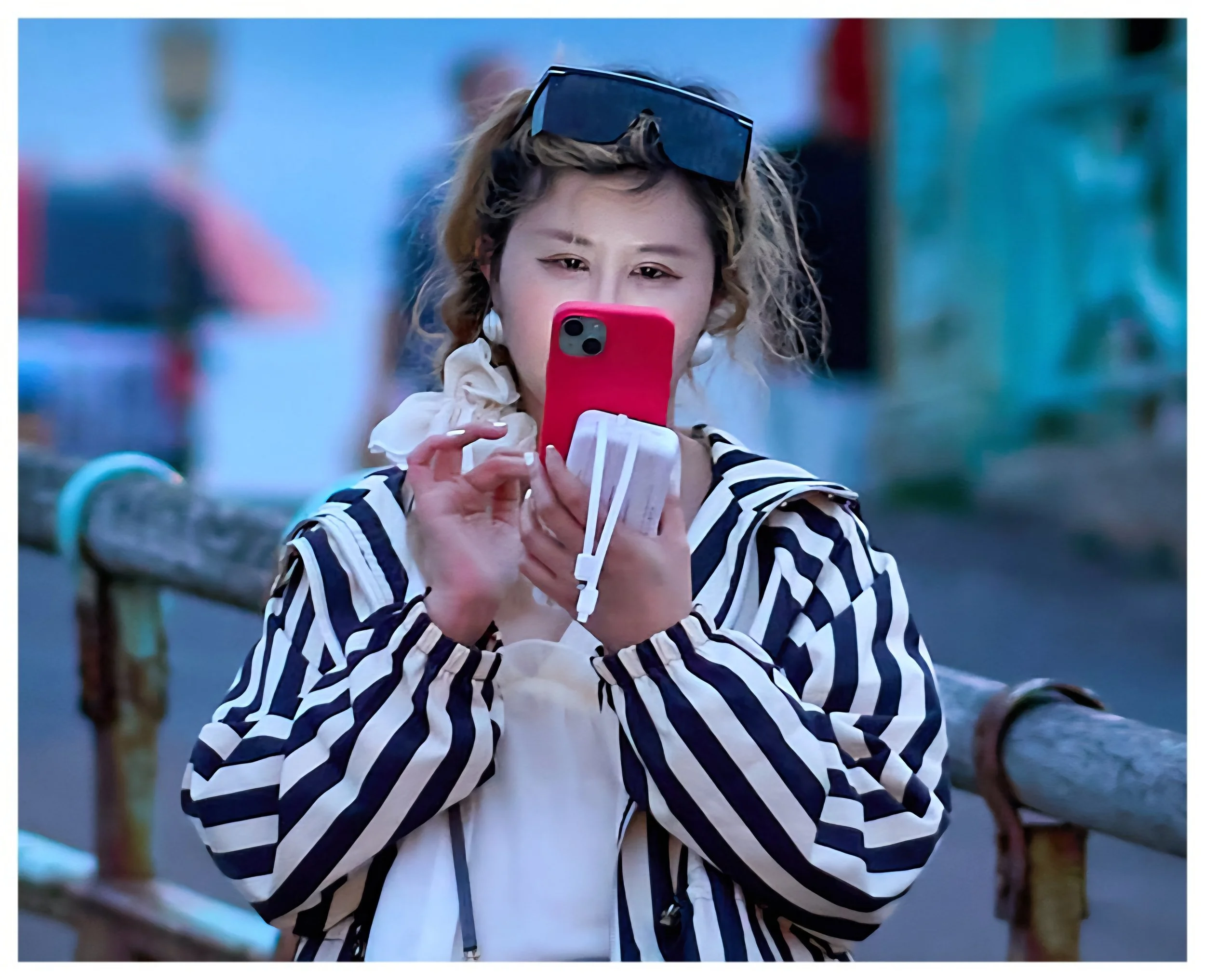 A woman with sunglasses on her head taking a photo with her pink phone while standing next to a green railing outside during the evening.