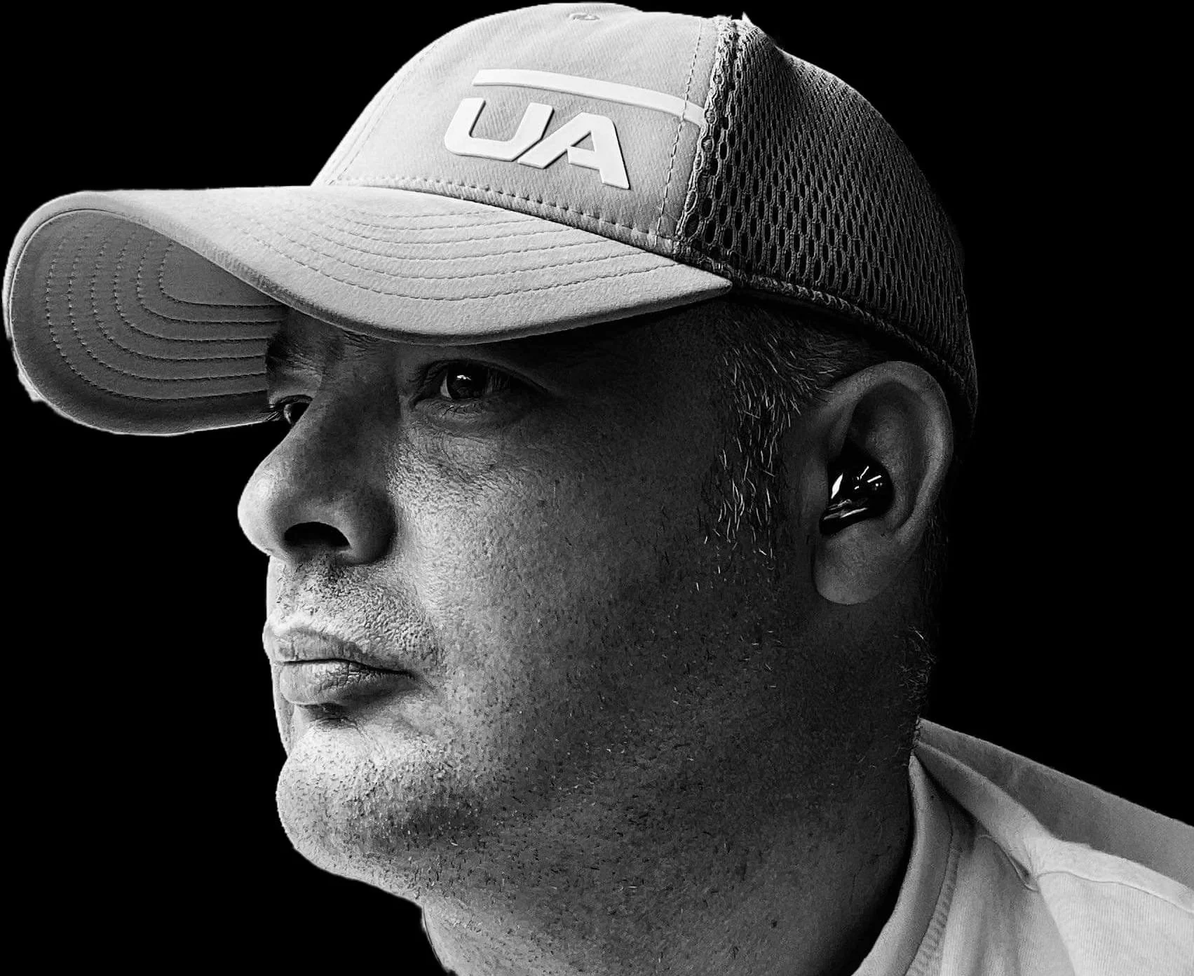 Black and white photograph of a man wearing a baseball cap with 'UA' logo, earbud in his ear, looking sideways with a serious expression.