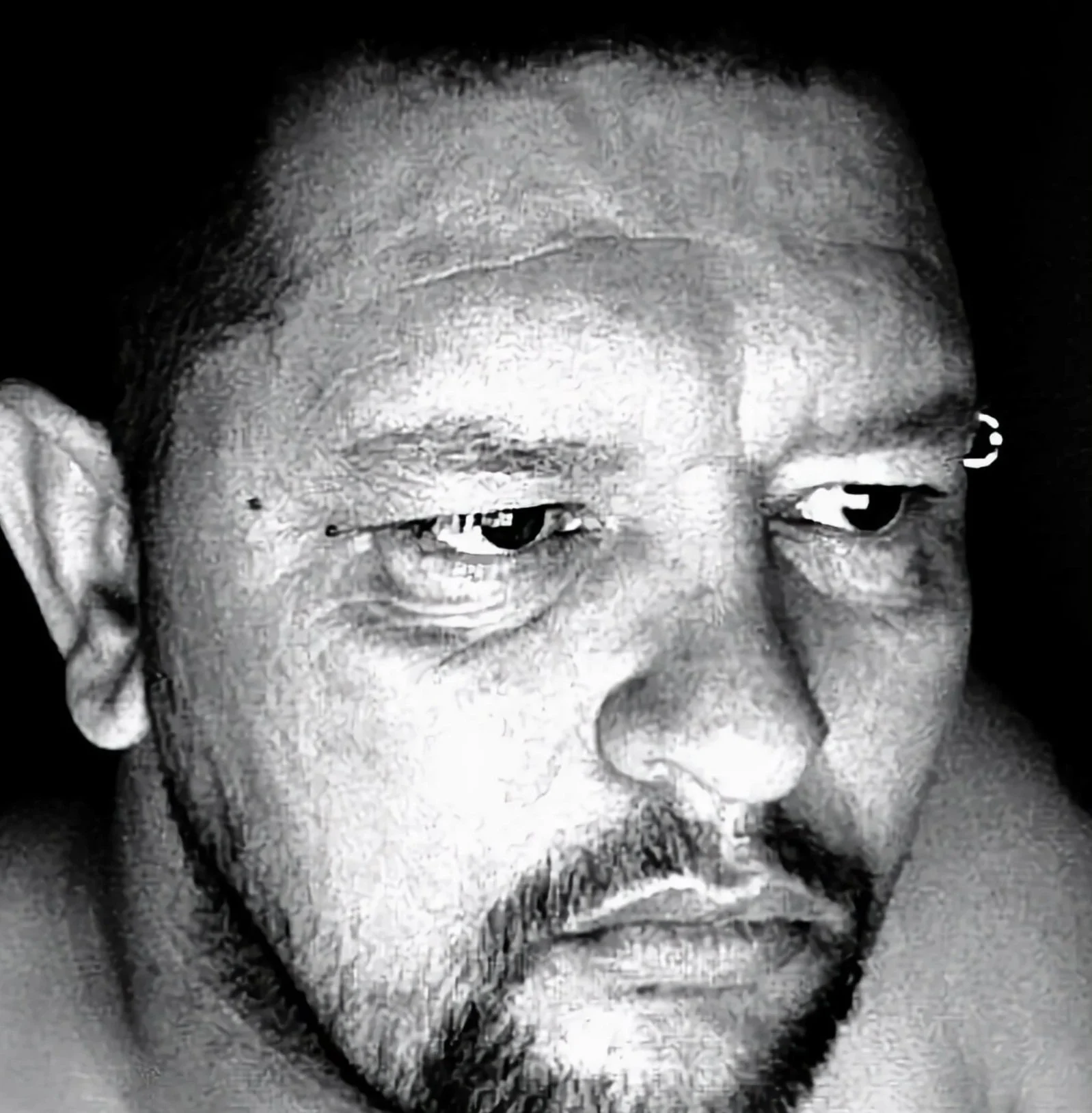 Close-up black-and-white photo of an Asian man's face with shaved head, beard, and mustache, looking down and to the side.