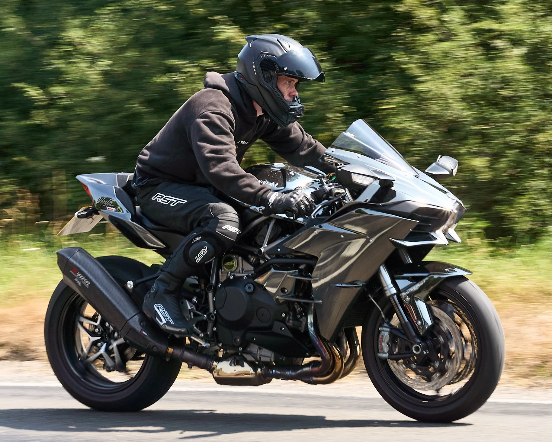 Motorcycle rider wearing a helmet and black protective gear riding a black motorcycle on a road with blurred green trees in the background.