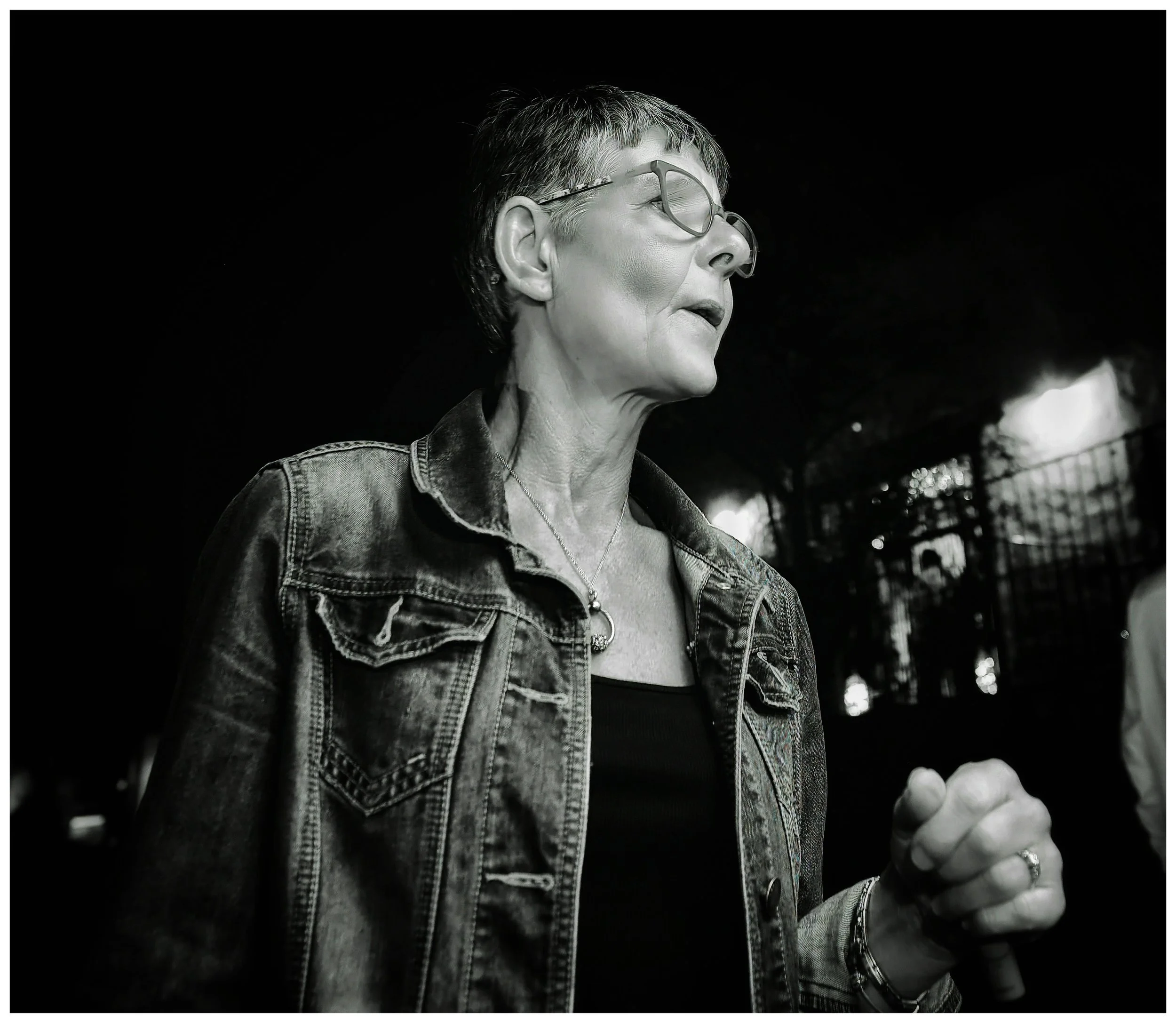 Black and white photo of an older woman with short hair and glasses, wearing a denim jacket and a necklace, speaking or singing into a microphone.