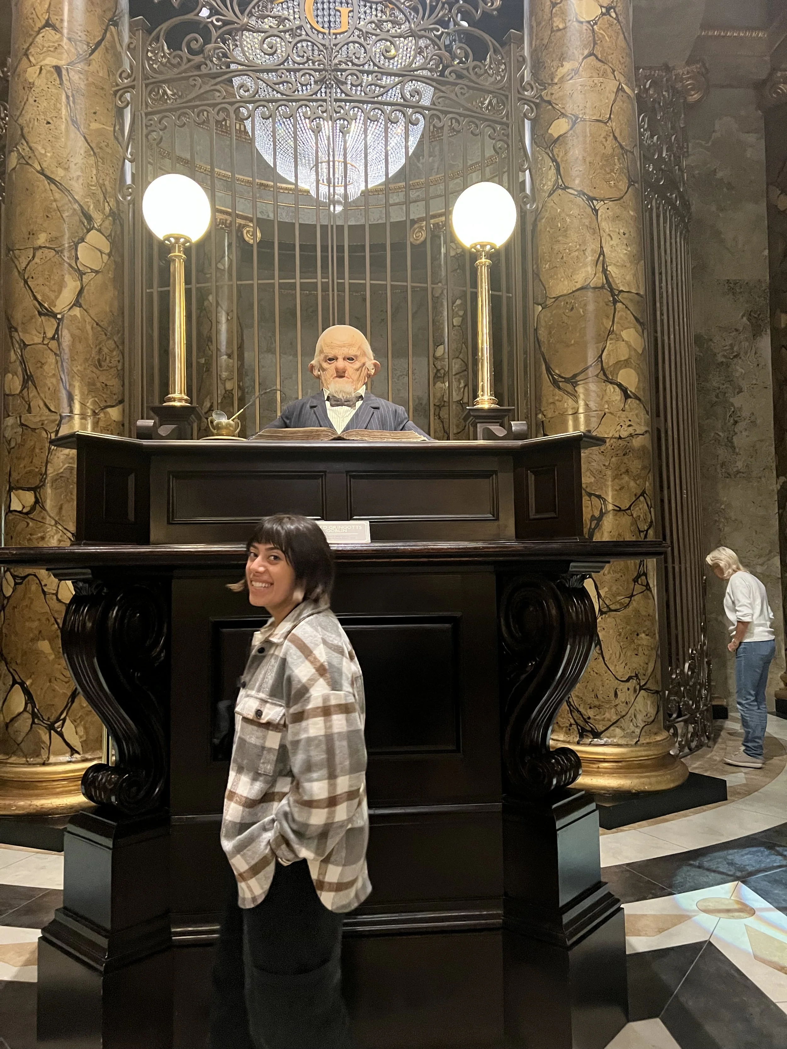 A woman in a plaid shirt is smiling and standing in front of a large ornate display case with a sculpture of an old man with a long face and bald head. The sculpture is behind a decorative iron gate, with two tall gold-colored lamps on either side and a chandelier above.