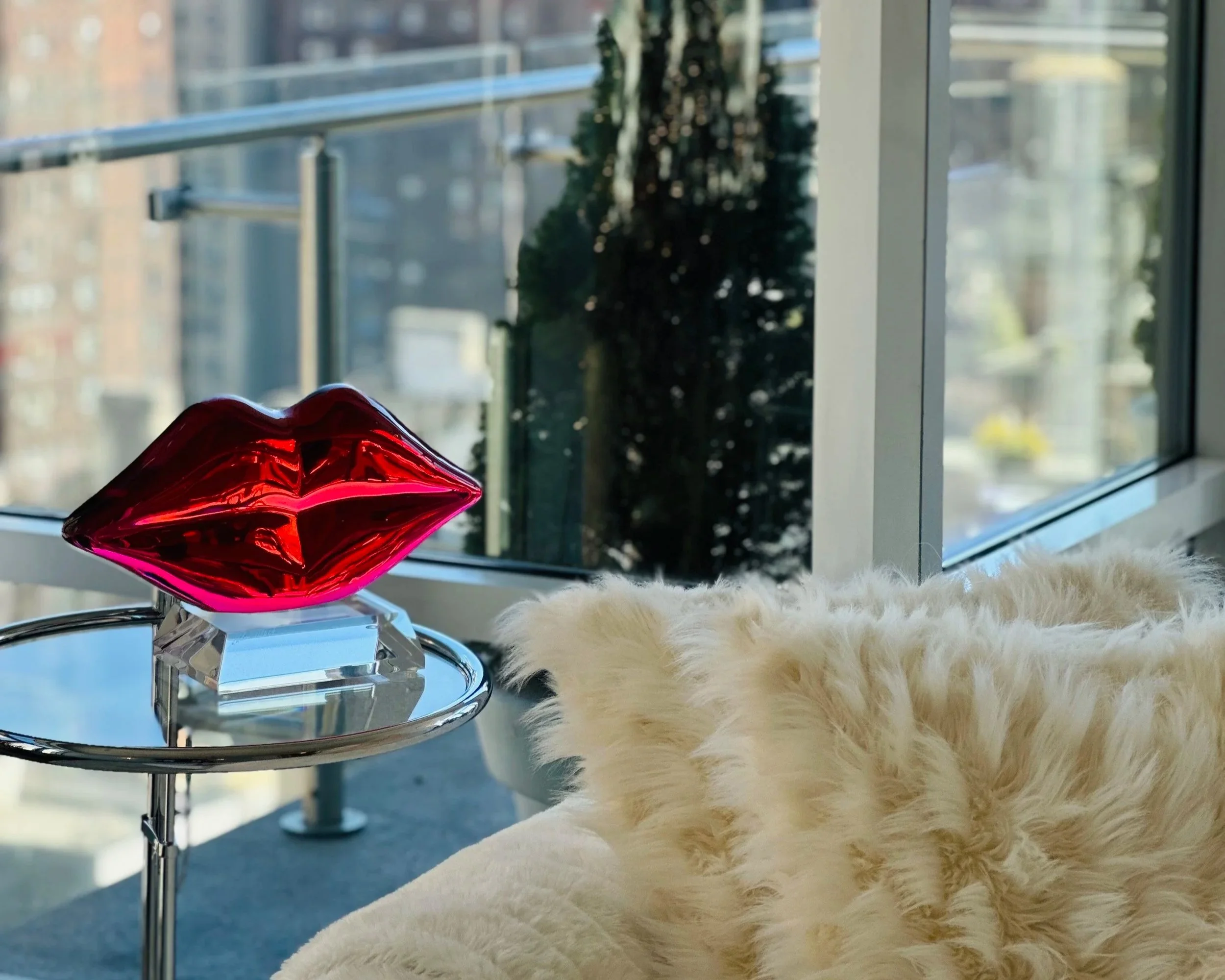 A decorative glass sculpture of red lips on a glass stand, placed on a round metal side table, next to a white, fluffy blanket or pillow in front of a large window with a cityscape view.