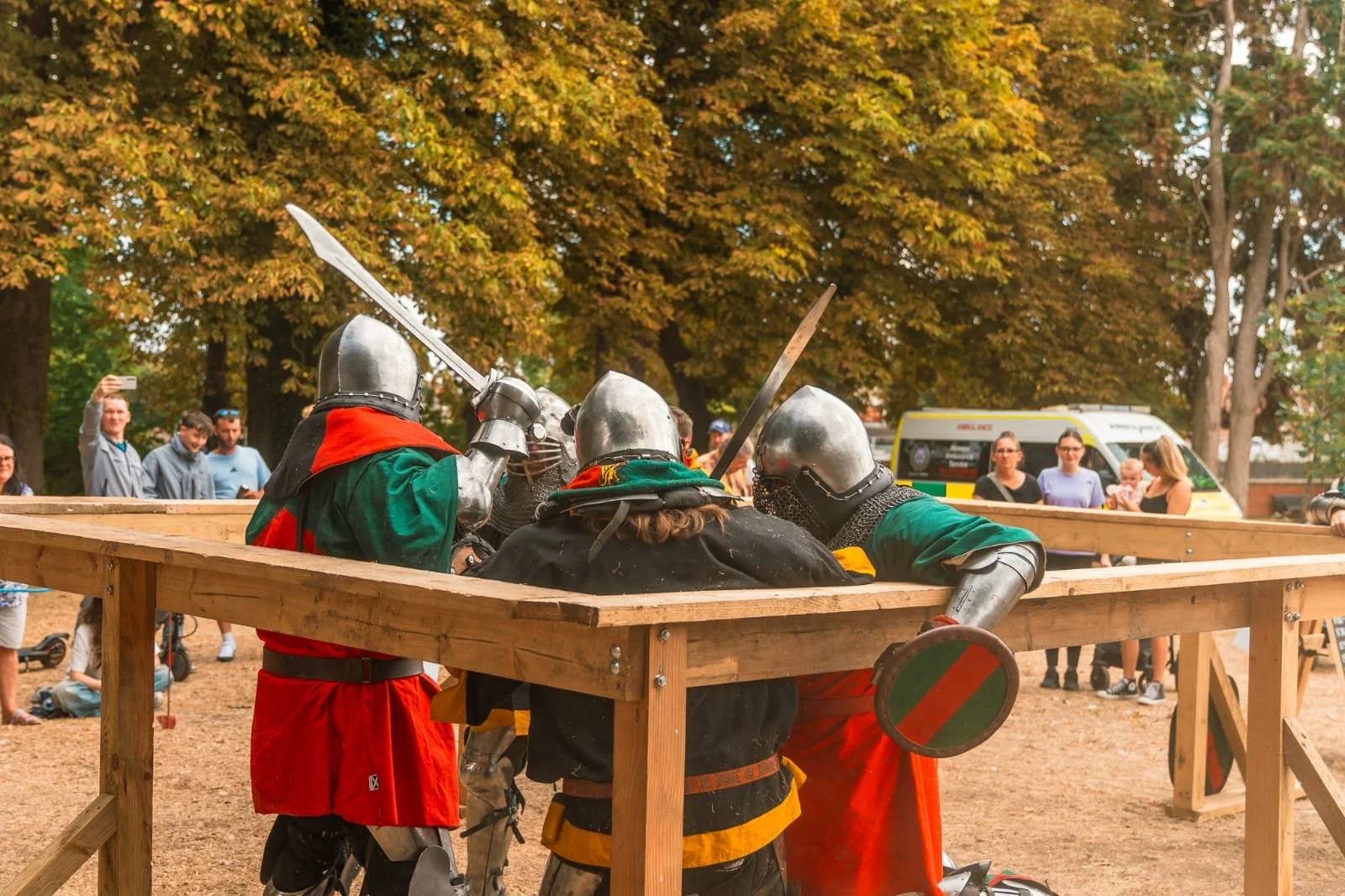 Four individuals dressed as medieval knights in armor battling with swords inside a wooden arena, with spectators watching nearby.