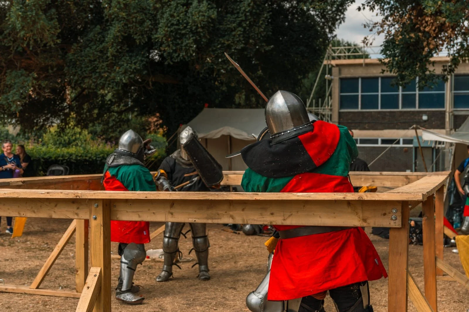 People dressed in medieval armor participating in a reenactment or event outdoors, with a wooden barrier in the foreground and trees and tents in the background.