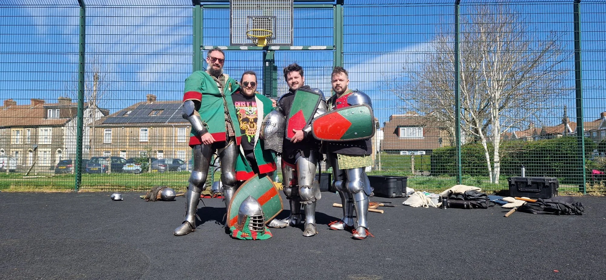 Four men dressed as medieval knights in armor, holding shields, standing in an outdoor sports court on a sunny day with houses and trees in the background.