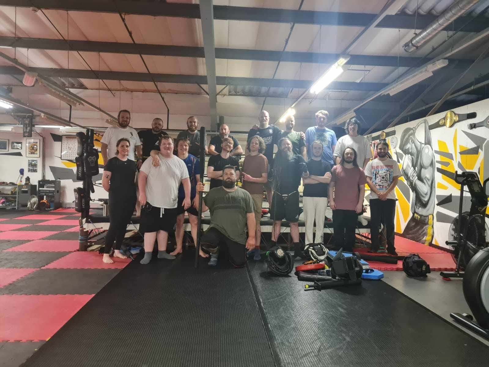 Group of people in a martial arts gym posing for a photo, with some on a raised platform and others on the floor, surrounded by gym equipment and colorful wall art.