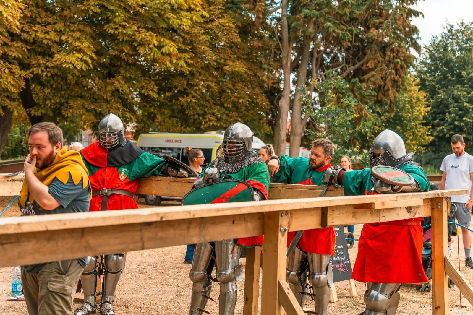 People dressed as medieval knights and jesters, with some wearing helmets and armor, standing behind a wooden barrier during an outdoor event.