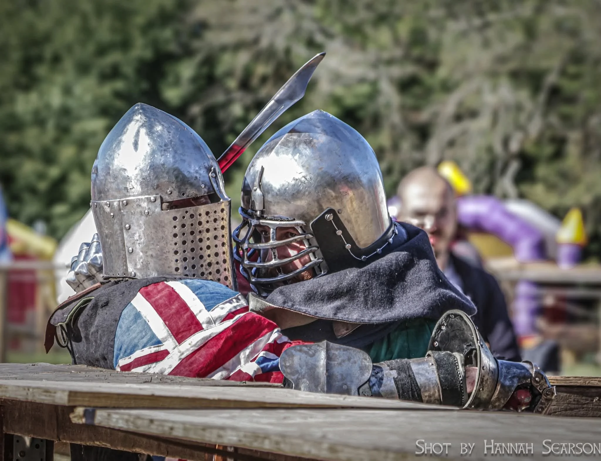 Two people in medieval armor engaged in mock combat at a fair or event, with a third person smiling in the background, outdoors with trees and tents.