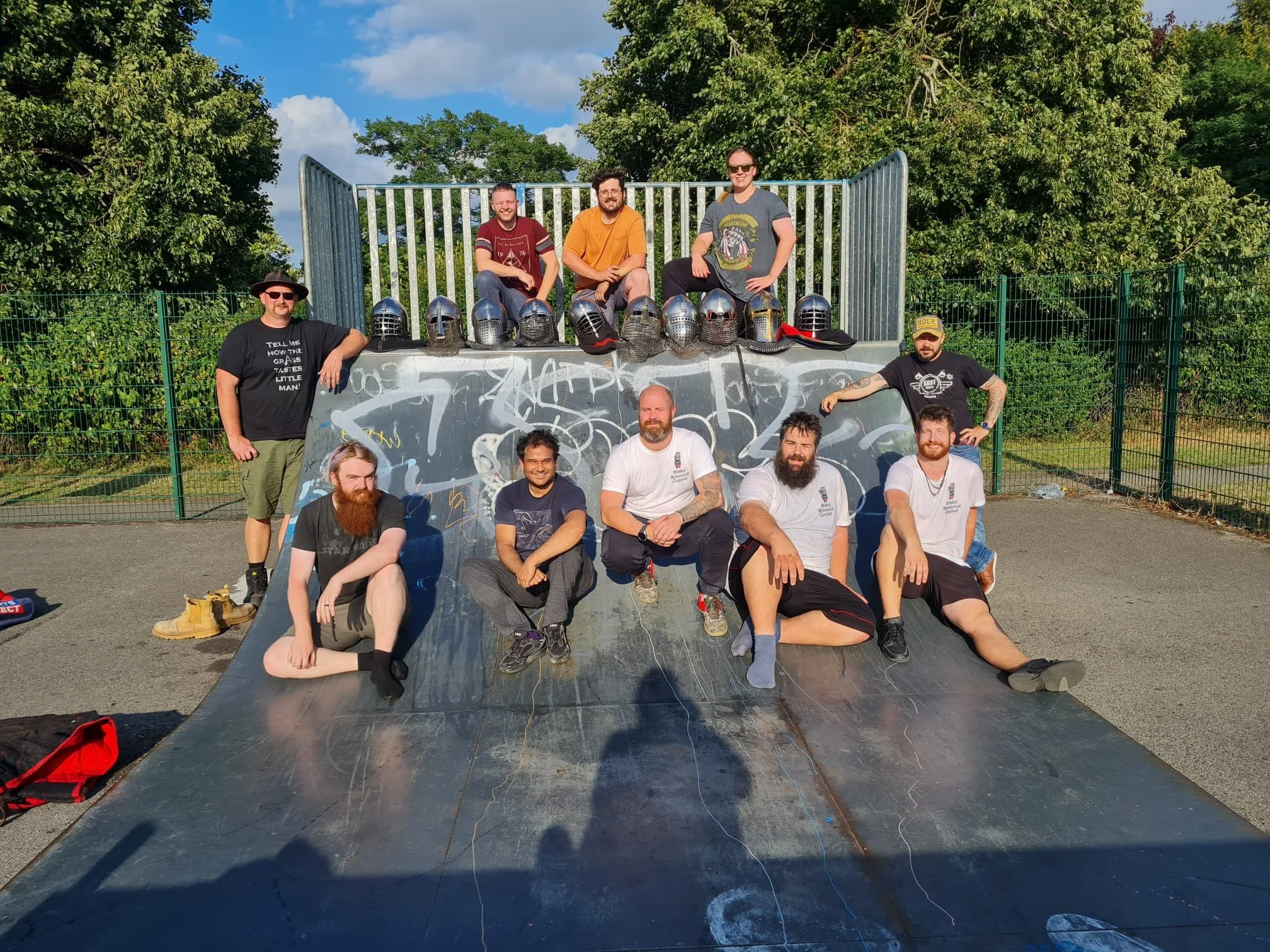 Group of ten friends at a skate park, posing together on a skate ramp with helmets lined up at the top, surrounded by green trees and blue sky.