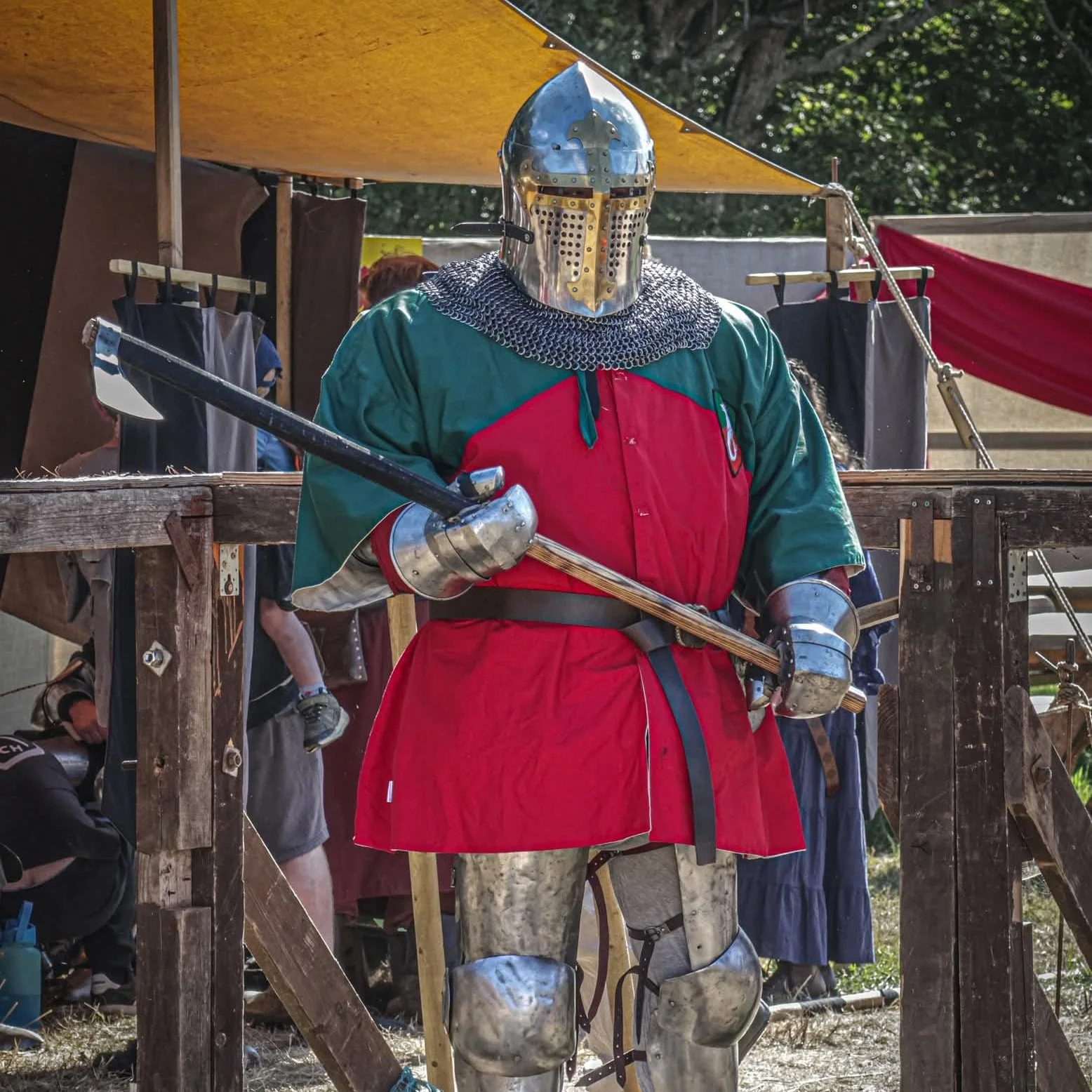 A person dressed as a medieval knight in armor, holding a sword, standing outdoors at a reenactment event or fair.