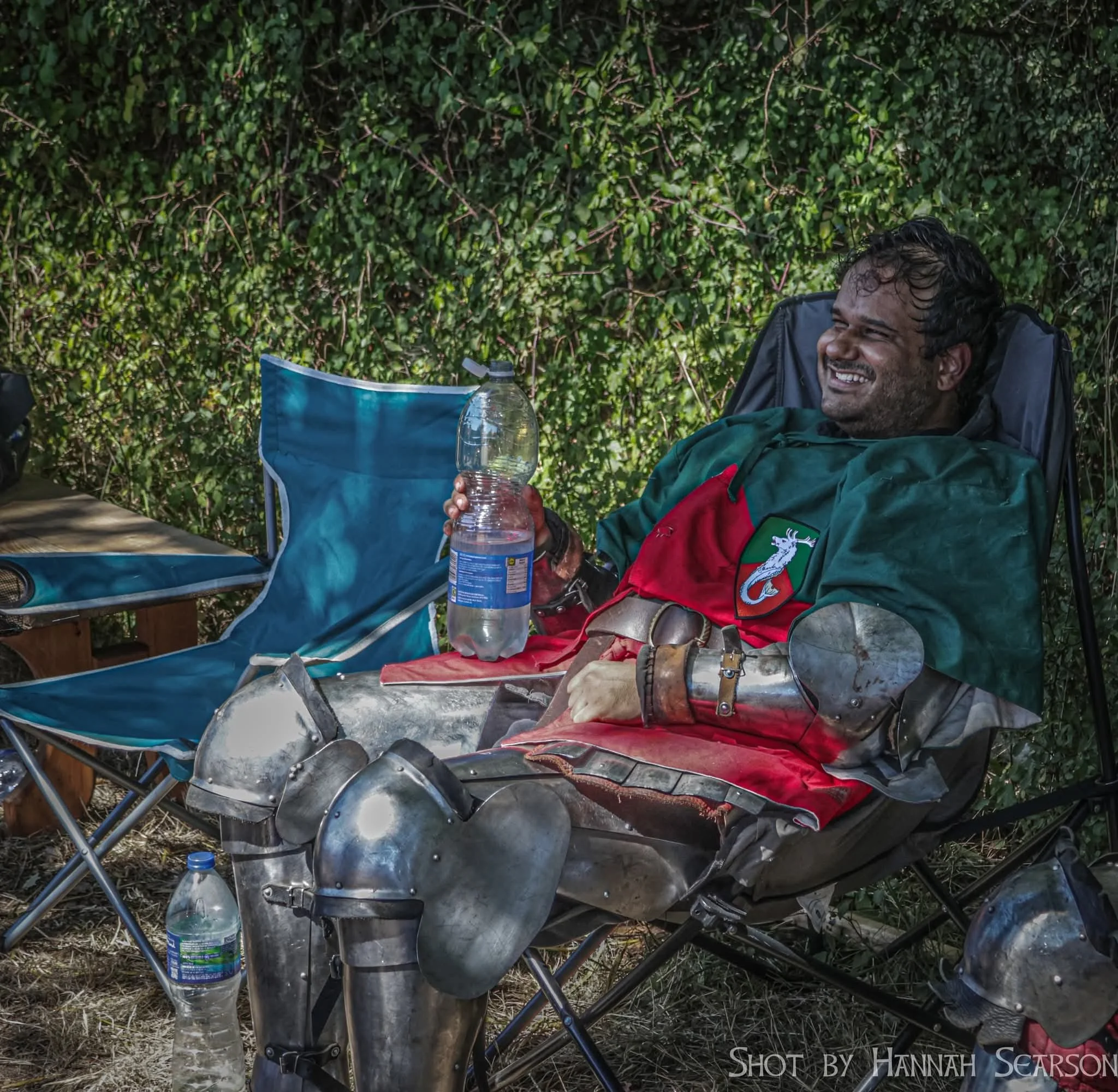 A man dressed as a medieval knight, wearing metal armor on legs and holding a plastic water bottle, sitting on a camping chair and smiling with a bushy green background.