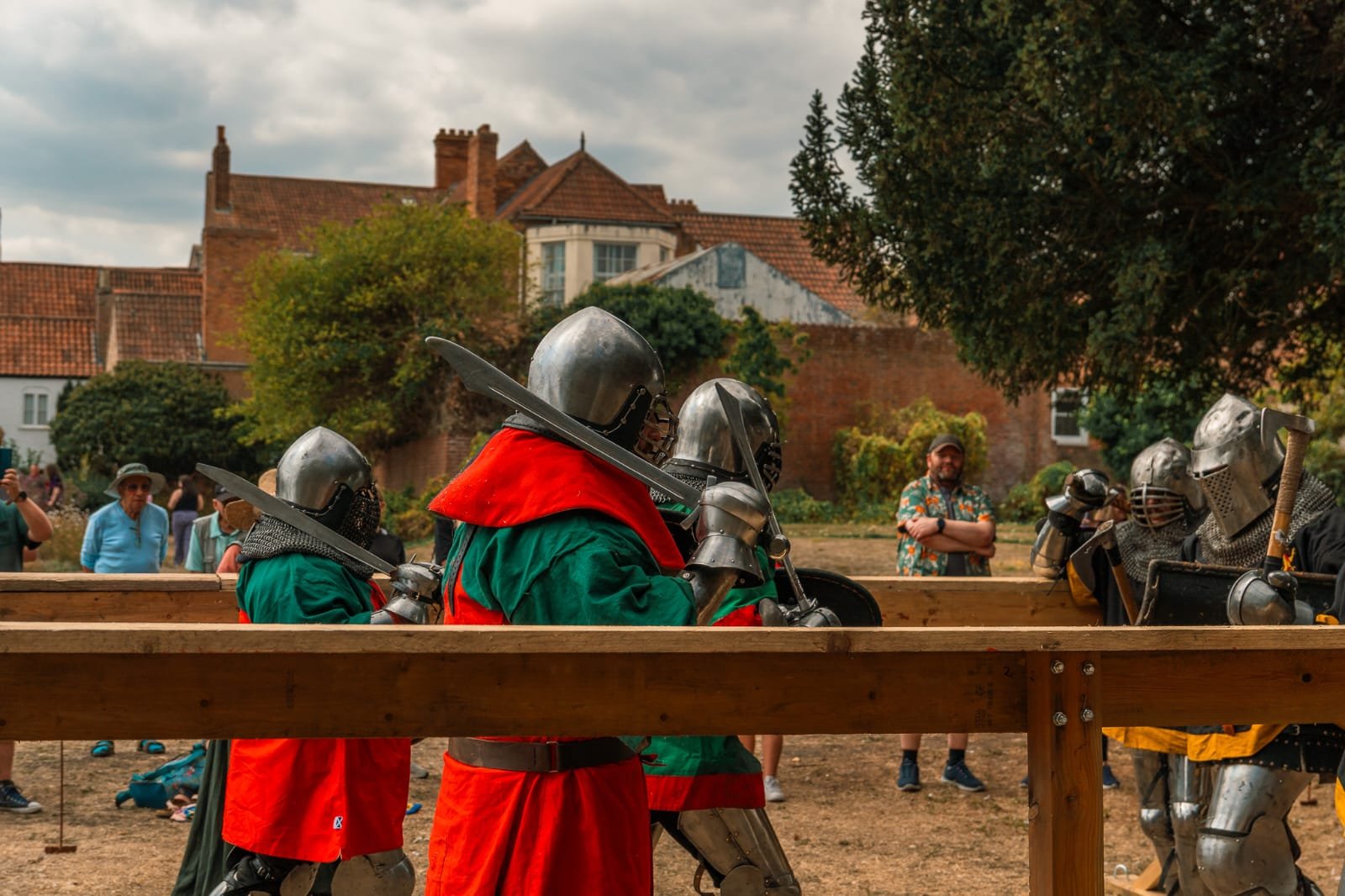 Group of people dressed as medieval knights with chainmail and helmets, practicing sword fighting or combat in a fenced arena outdoors, with onlookers watching.