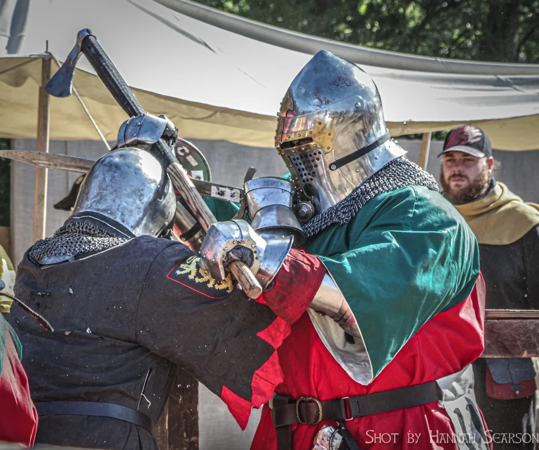 Two men in medieval knight armor are engaged in a sword fight during a reenactment event, with a third man observing in the background.