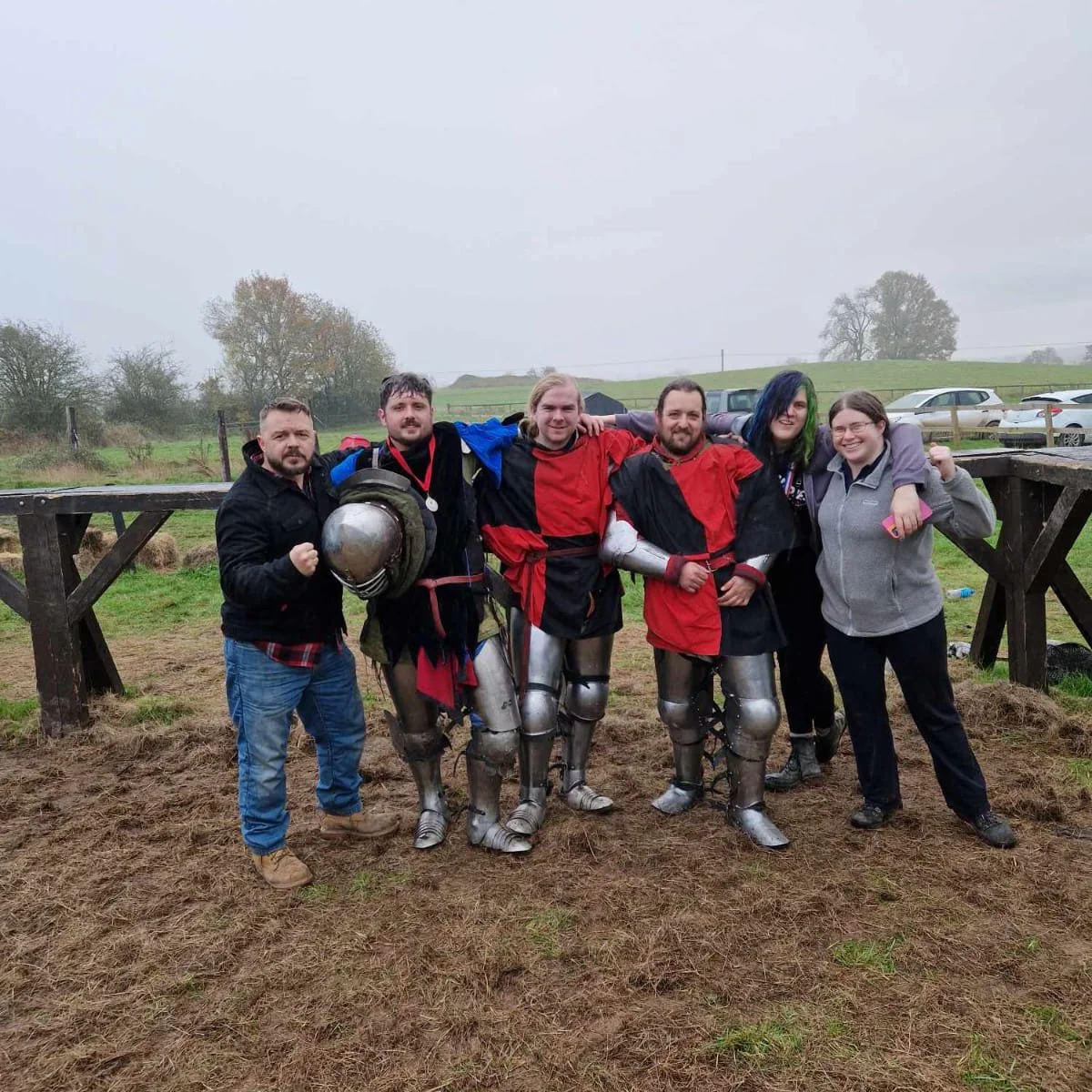Group of five people standing outdoors on muddy ground, with some wearing medieval-style knight armor and others in casual clothing, celebrating or posing after a game or event.