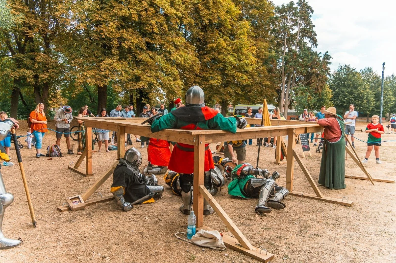 People watching a medieval knight demonstration with statues of knights in armor, set in a park on a sunny day