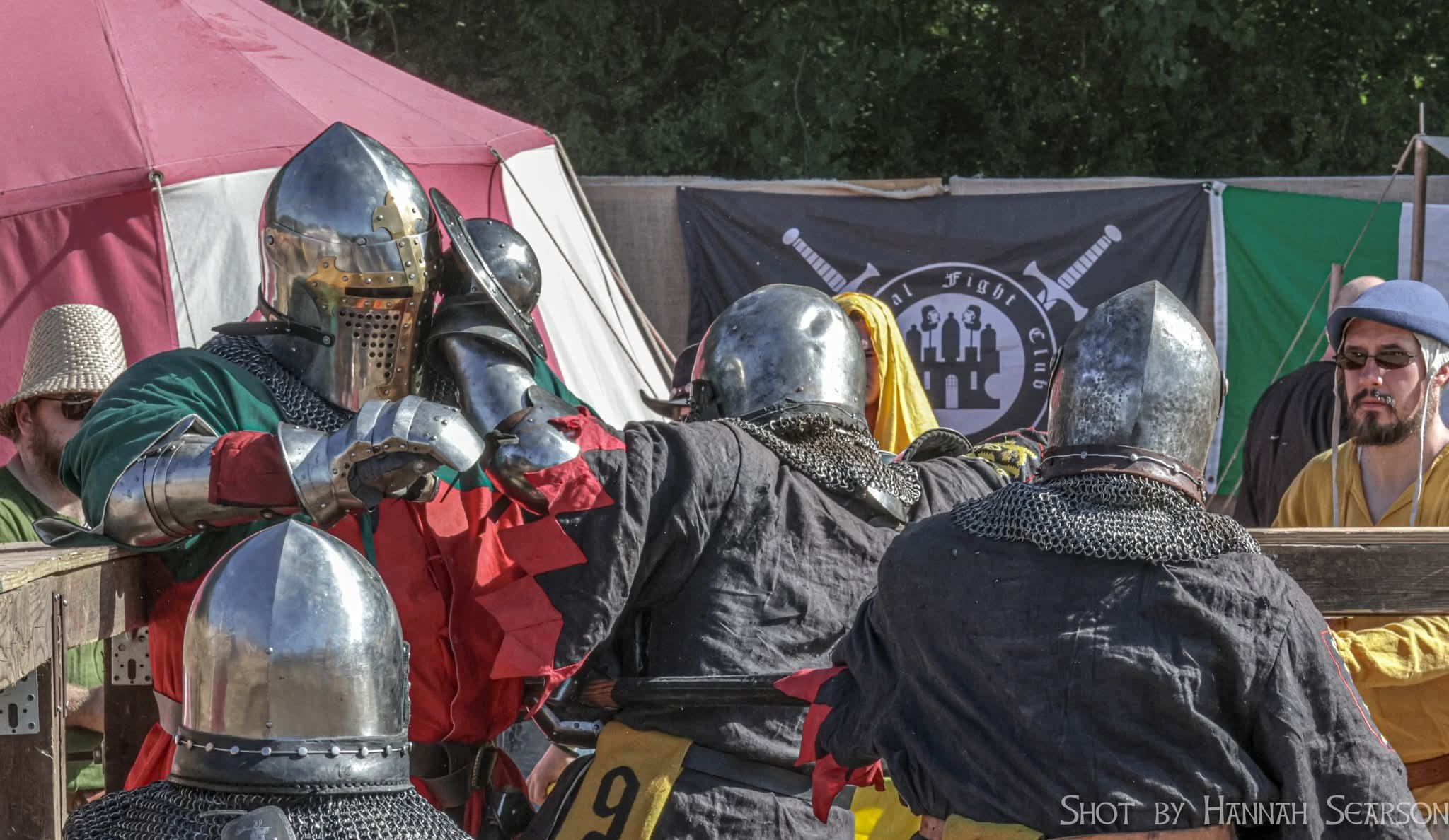 Four medieval knights in armor and costumes are engaging in a mock combat at a renaissance fair or reenactment event. Behind them, spectators and a banner with swords and a coat of arms are visible.