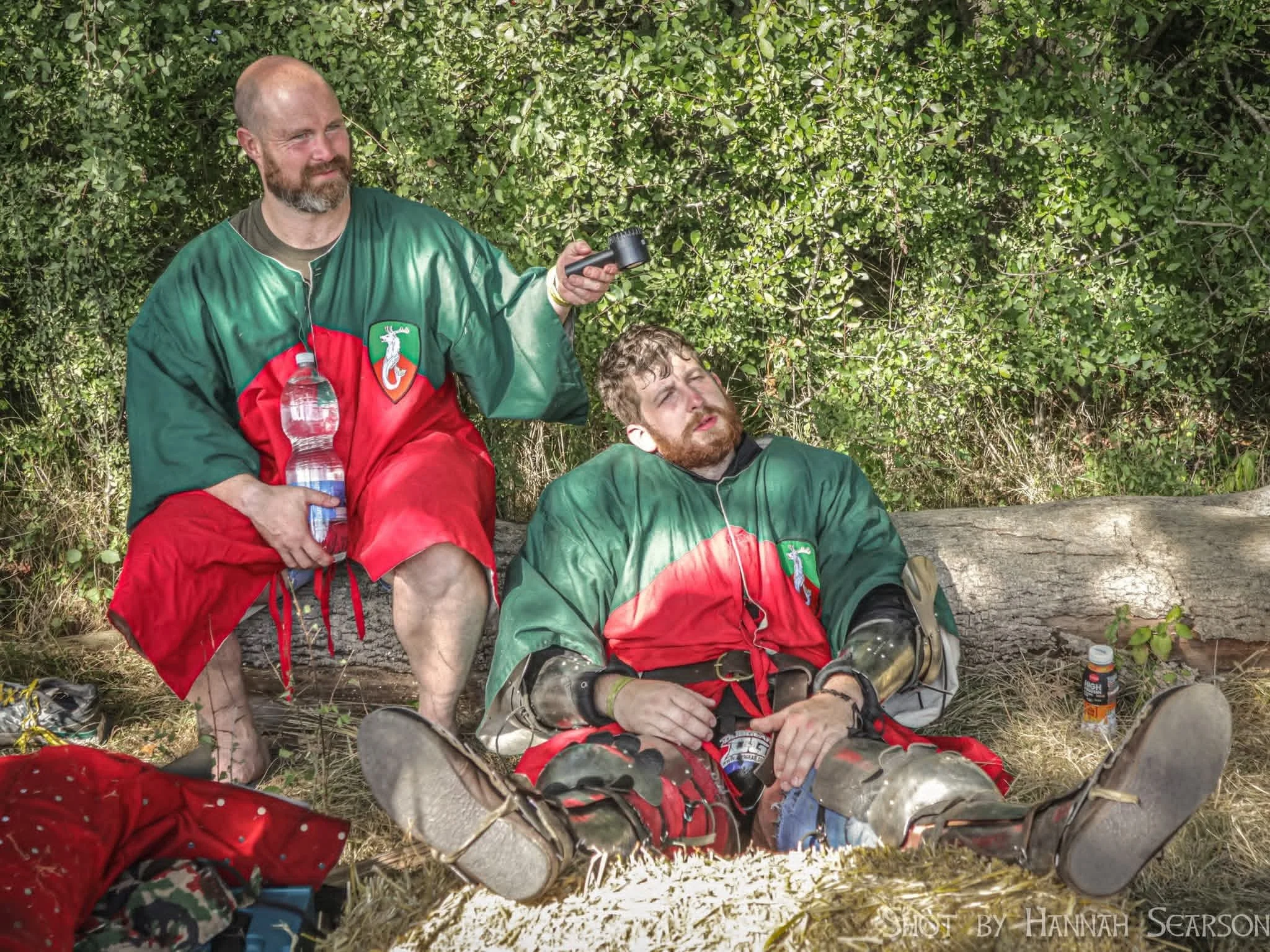 Two men in hockey jerseys outdoors, one sitting and resting with eyes closed, and the other standing with a water bottle and phone, surrounded by greenery and camping gear.