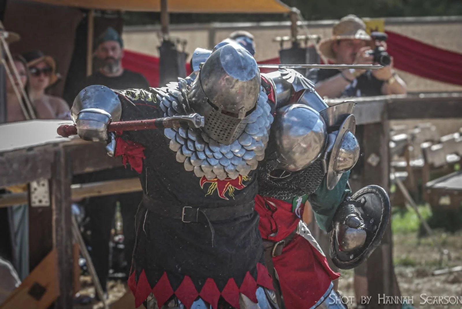 A person dressed as a medieval knight in chainmail armor and a helmet, engaging in a sword fight at a Renaissance fair or medieval reenactment event with spectators in the background.