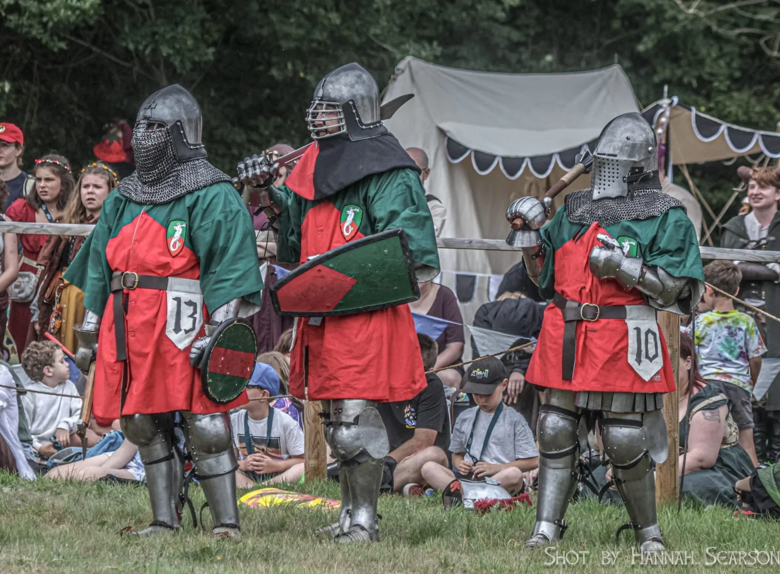 Three individuals dressed in medieval knight costumes, including armor and helmets, standing on grass with spectators and tents in the background.