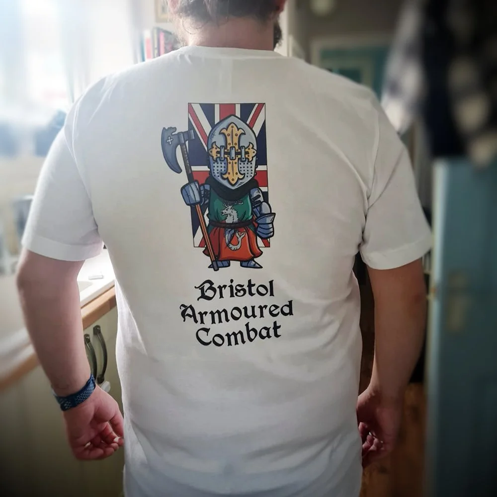 Person wearing a white T-shirt with a cartoon knight in armor, holding an axe, with a background of the Union Jack flag, and the text 'Bristol Armoured Combat' printed below.