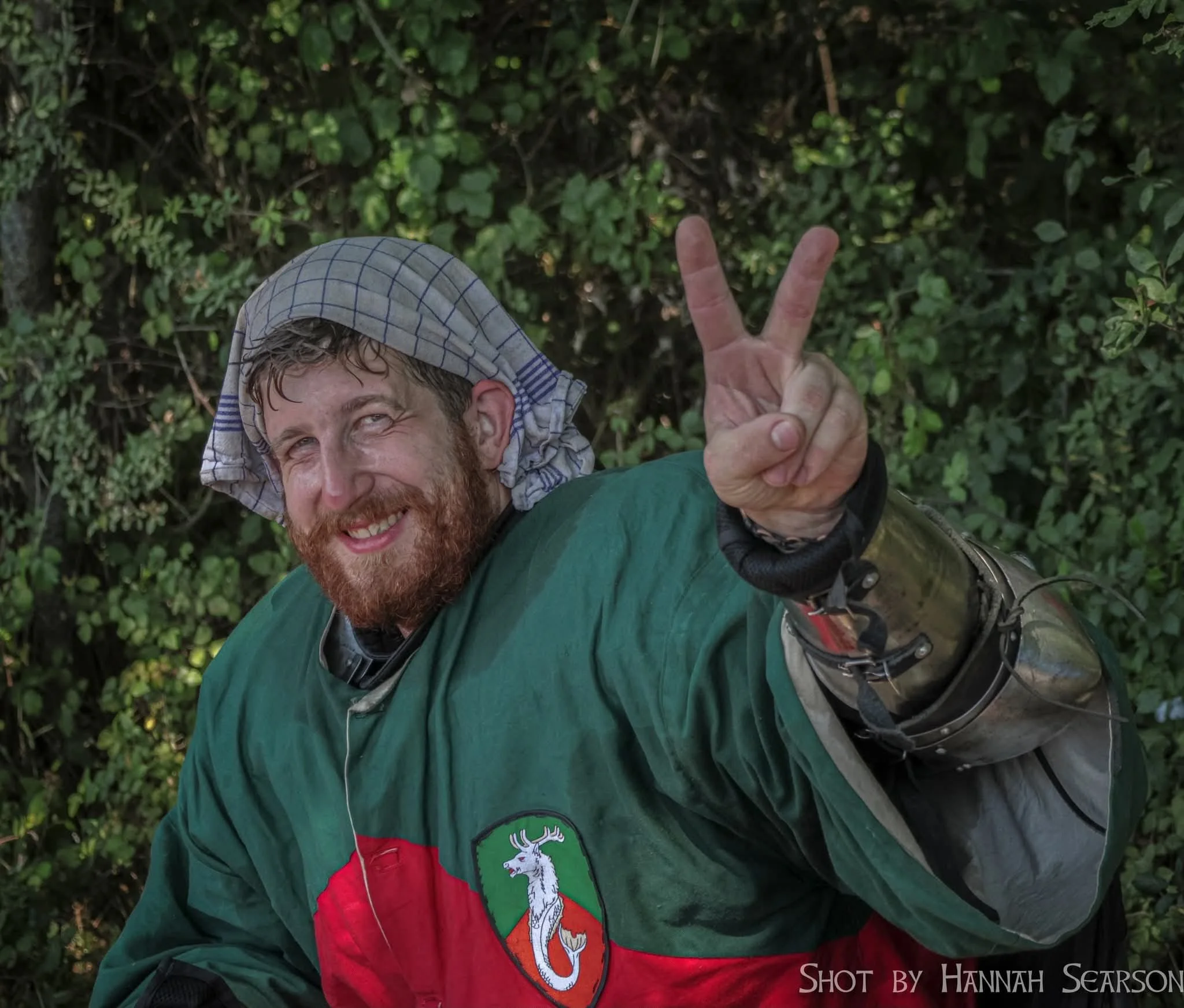 A smiling man with a beard and a plaid cloth on his head, wearing a green and red jacket with a crest of a white deer on a green shield, giving a peace sign with his right hand, standing in front of a leafy green background.