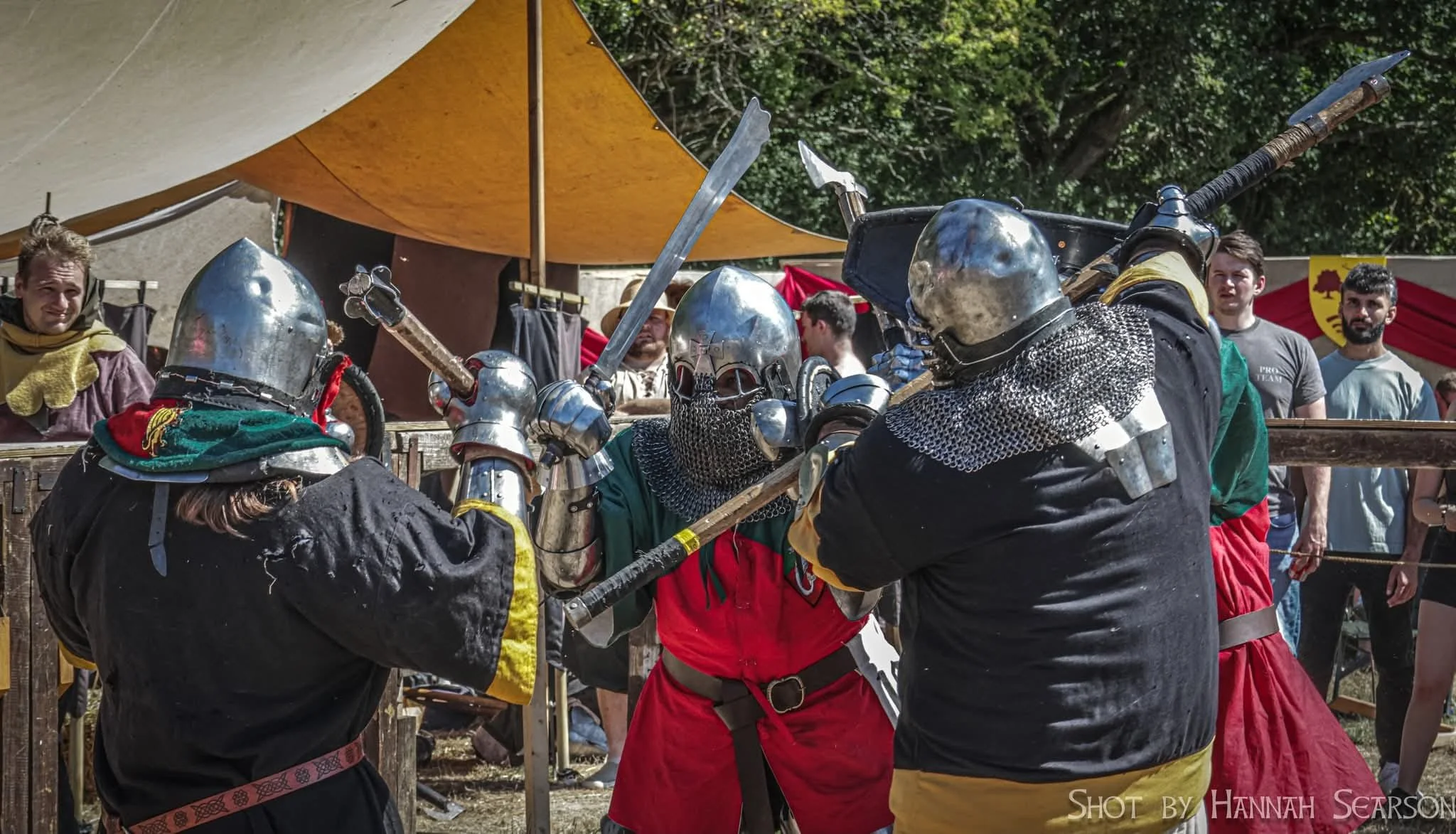 Reenactment of medieval combat with four knights in armor fighting with swords at a historical fair, with spectators in medieval and casual clothing watching.