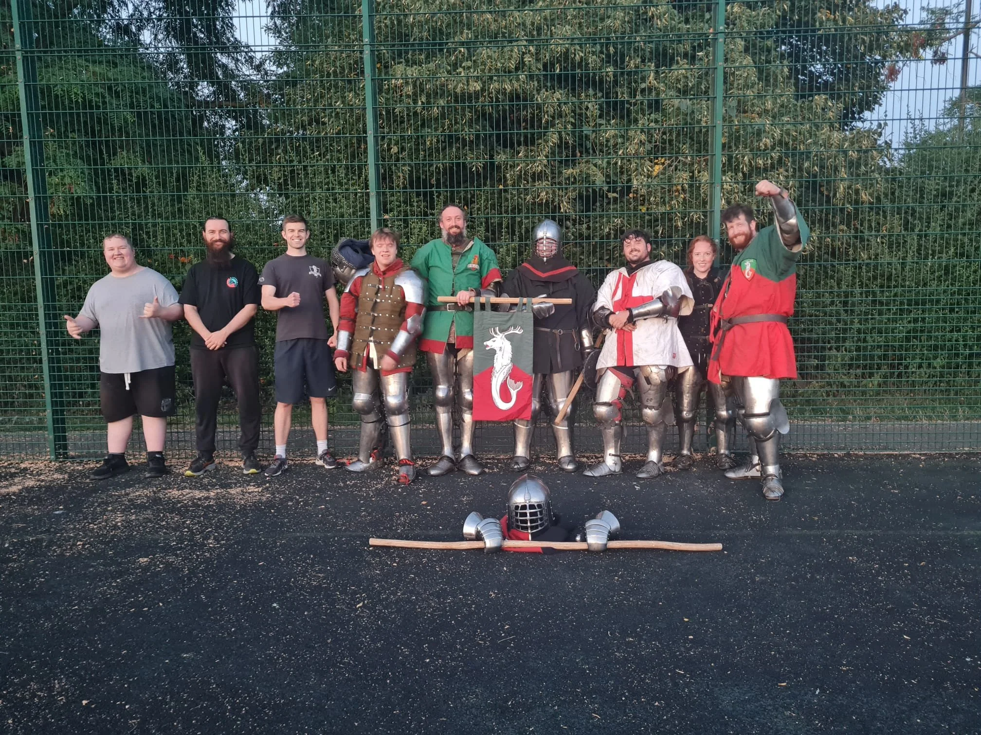 A group of nine people, some in medieval armor and costumes, standing in front of a green metal fence, posing for a photo on a blacktop outdoor surface.