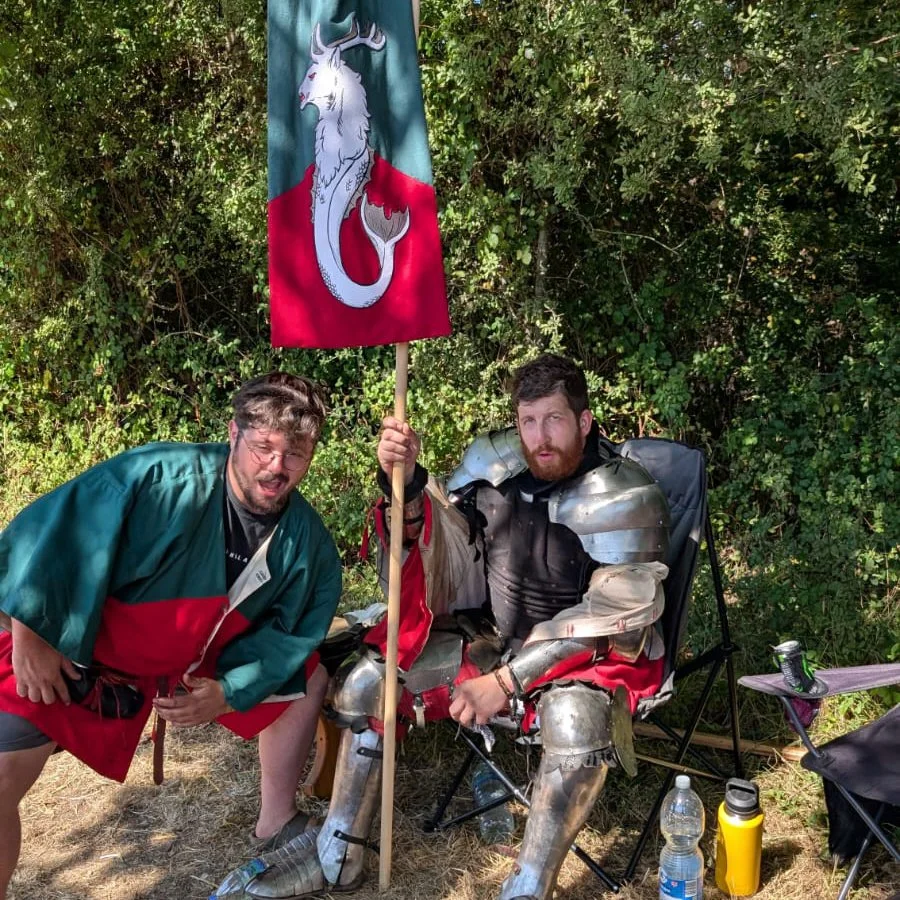 Two men dressed in medieval armor and clothing, one sitting in a camp chair with armor and holding a flag with a sword-wielding unicorn illustration, outdoors with trees in the background.