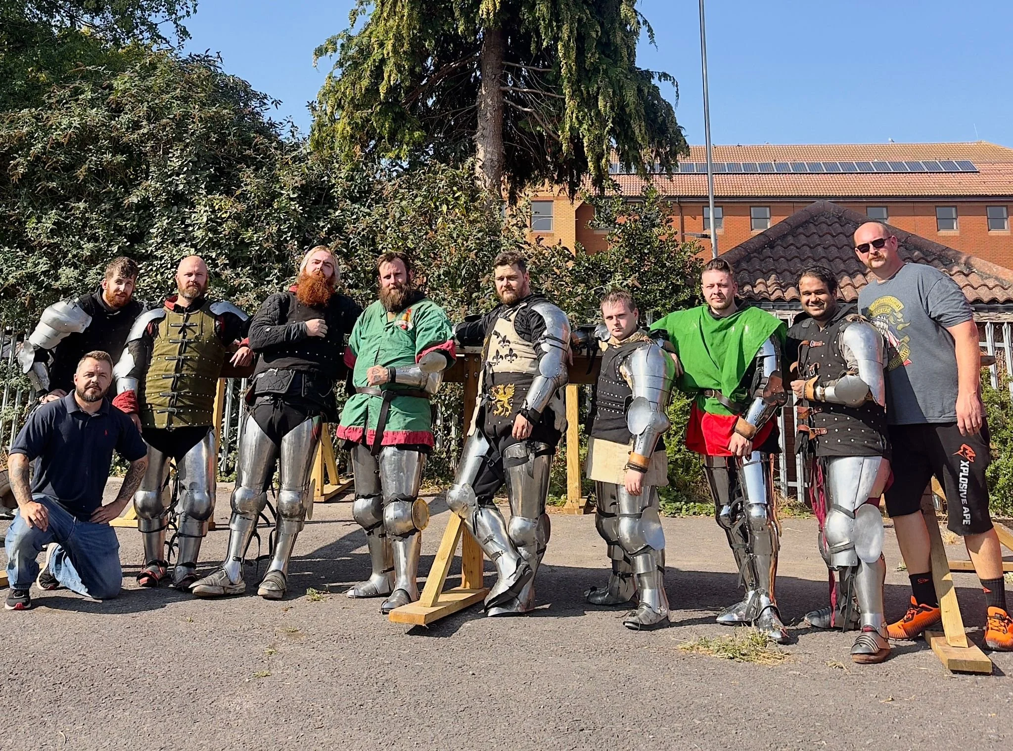 A group of people dressed in medieval armor, with some in swords and chainmail, posing outdoors on a sunny day. There are ten men, some standing and one kneeling, with green trees and a building with a tiled roof in the background.