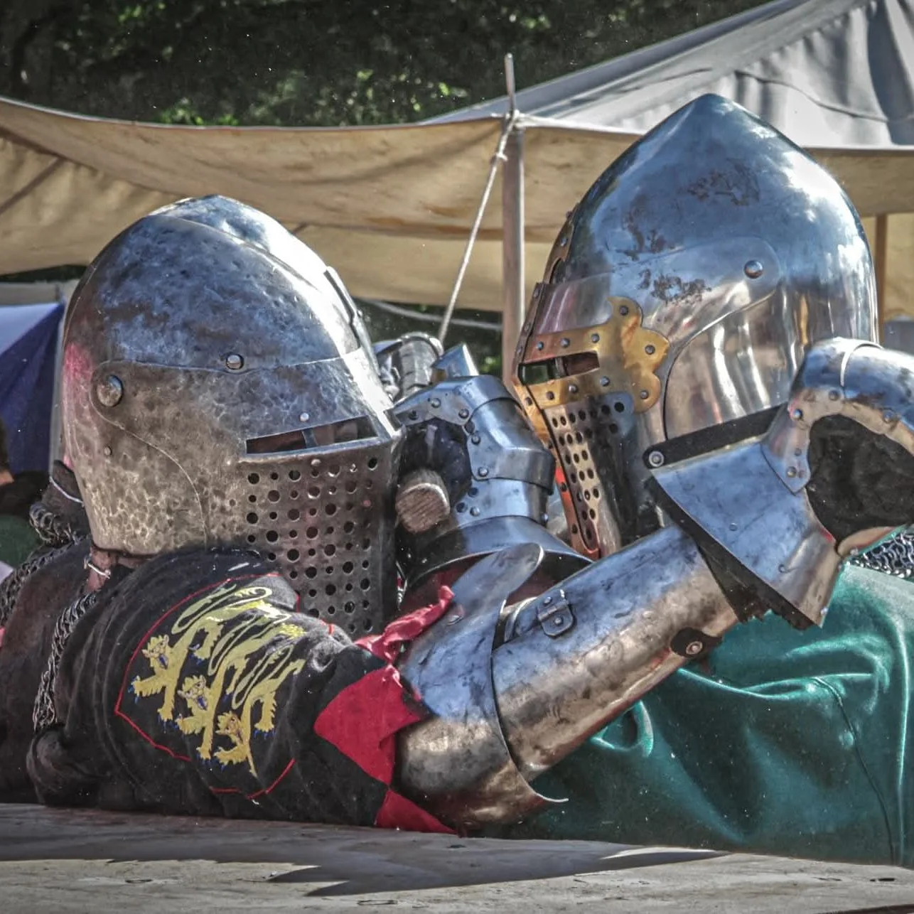 Two knights wearing metal helmets and armor dueling with swords during a reenactment or event.