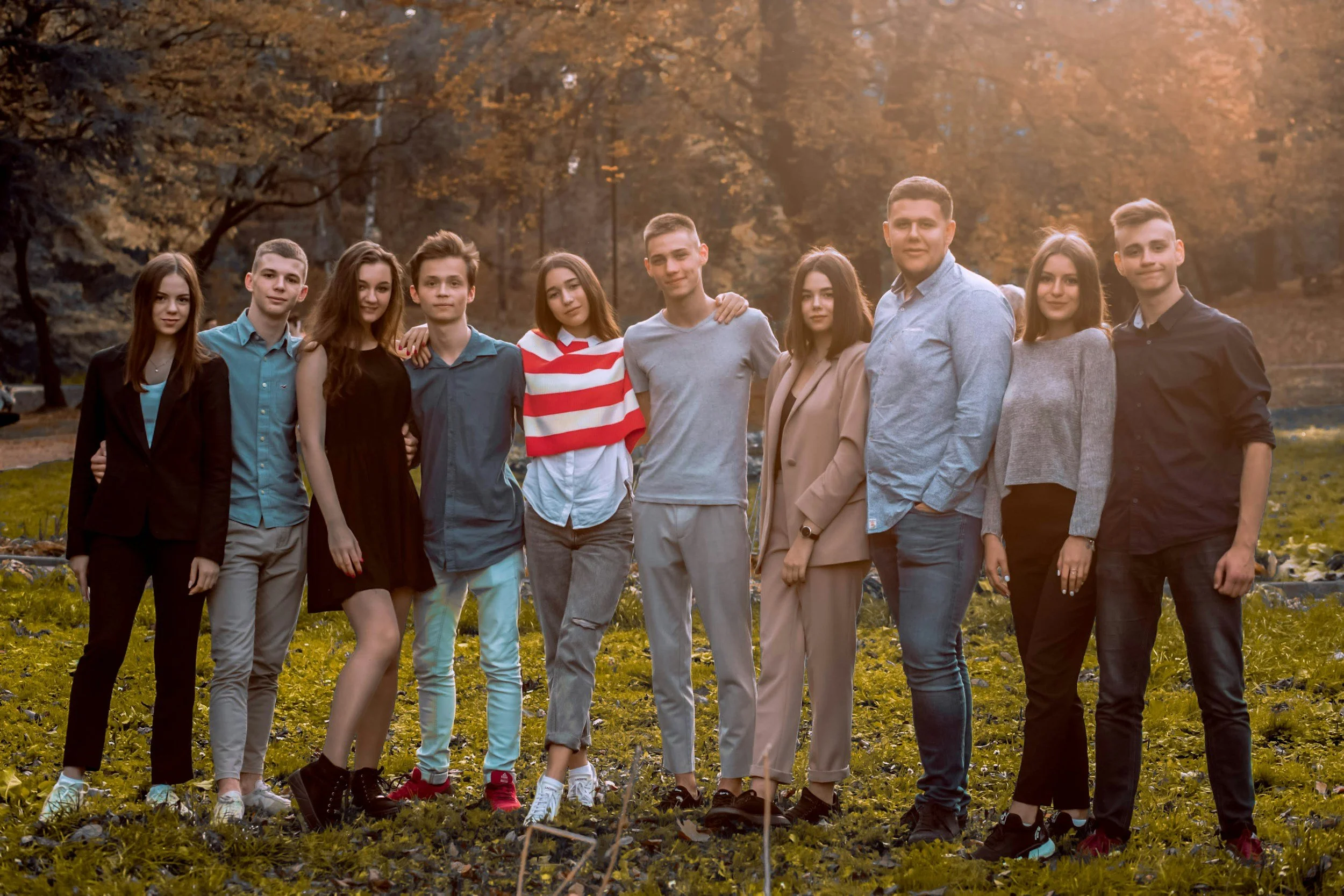A group of family members at a reunion  standing outdoors on grass during autumn, with trees and sunlight in the background.