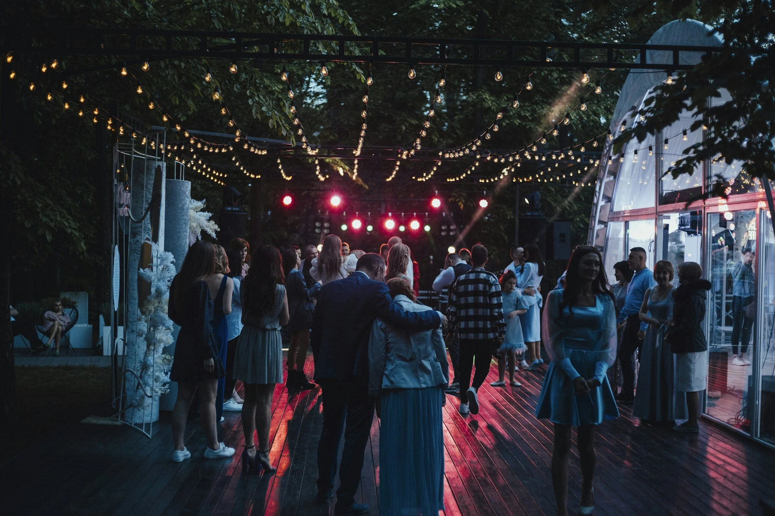 People dancing and socializing at an outdoor event during the evening, with string lights overhead and colorful stage lights in the background.