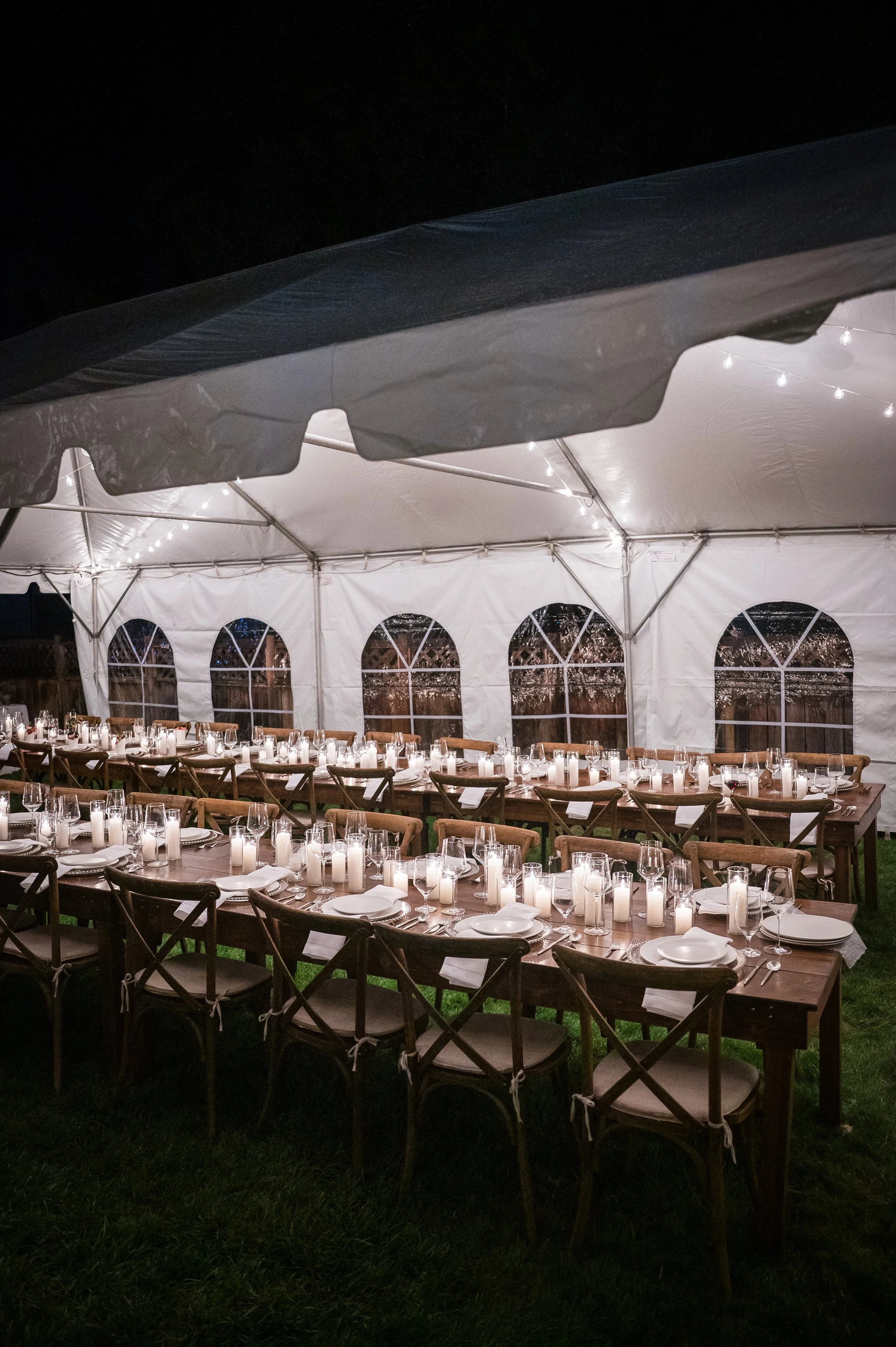 A large white event tent and banquet tables set up outdoors on green grass with farm equipment, including red tractors and farming tools, visible through the tent. The sky is partly cloudy with patches of blue.