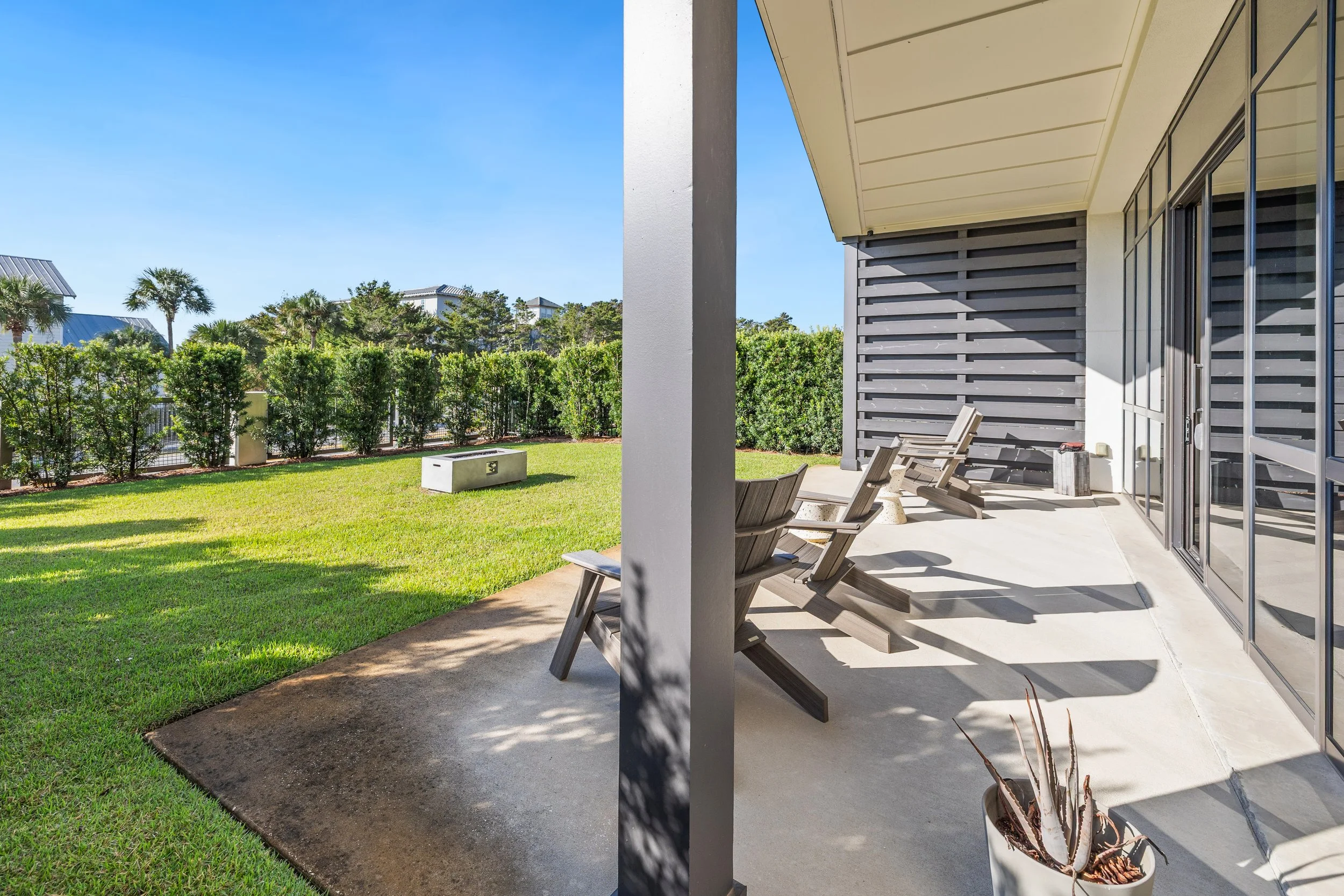 Patio with wooden chairs, potted plant, and sliding glass doors, overlooking a grassy yard enclosed by a fence hedge under a clear blue sky.