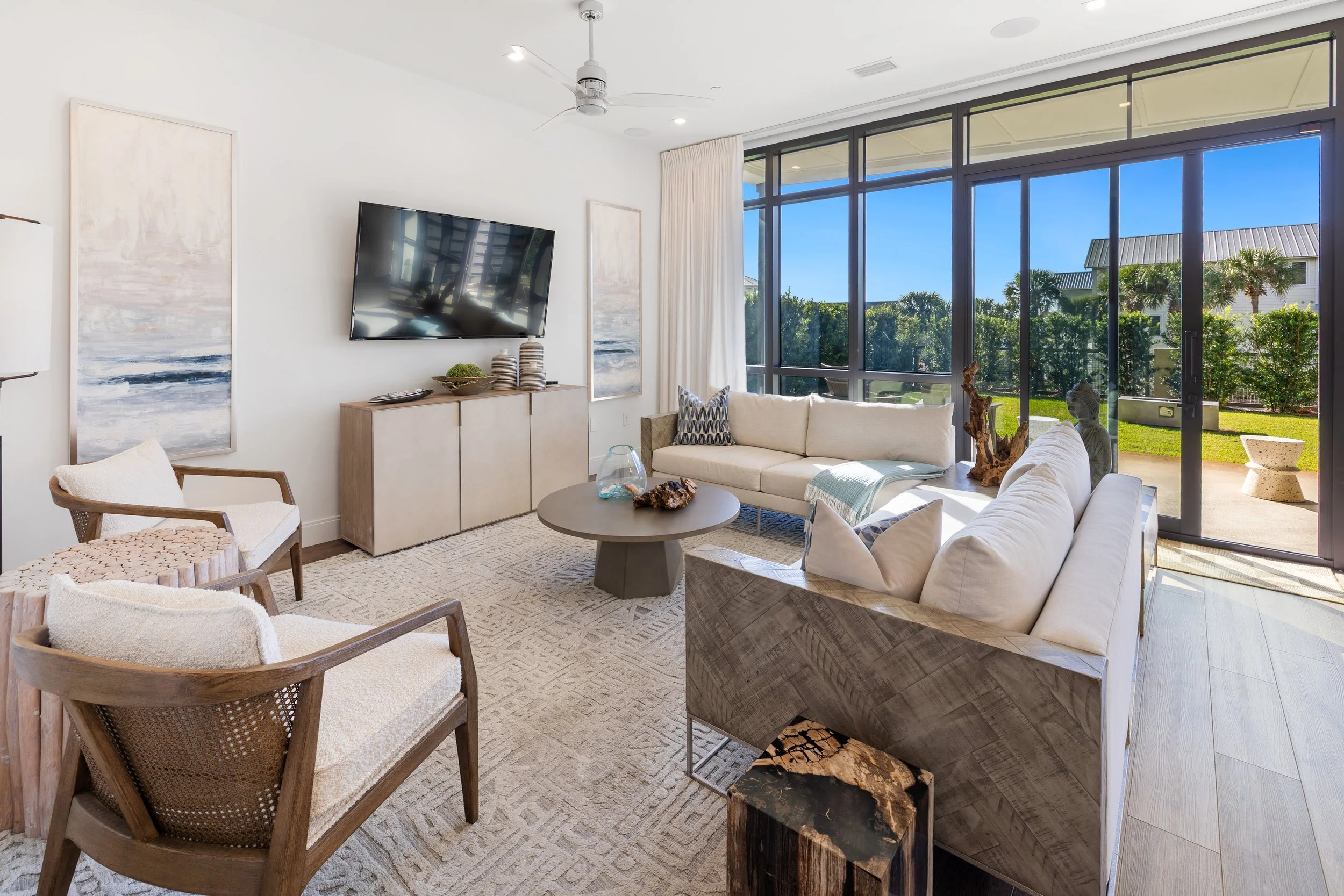Bright living room with large glass sliding doors showing outdoor greenery, featuring white sofas, wooden chairs with woven seats, a round coffee table, a TV and modern art decor.