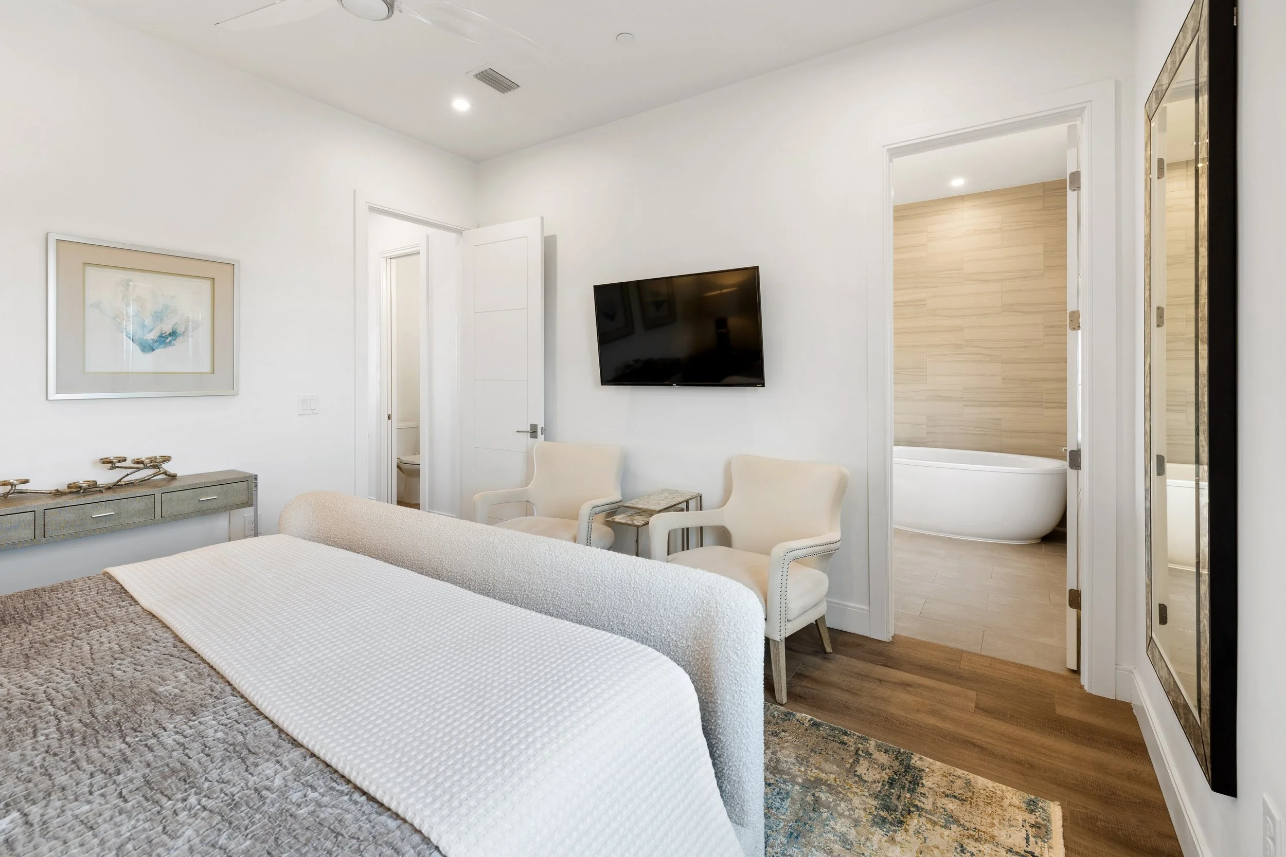 Modern master bedroom with a king bed, two white armchairs, a side table, wall-mounted TV, bathroom with a soaking tub.