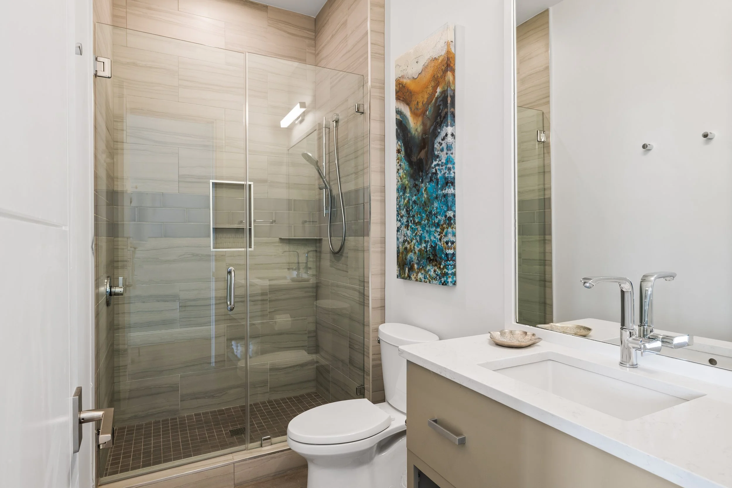First floor modern bathroom with a glass-enclosed shower, a white toilet, a beige vanity with a sink, a large mirror, and a colorful abstract painting on the wall.