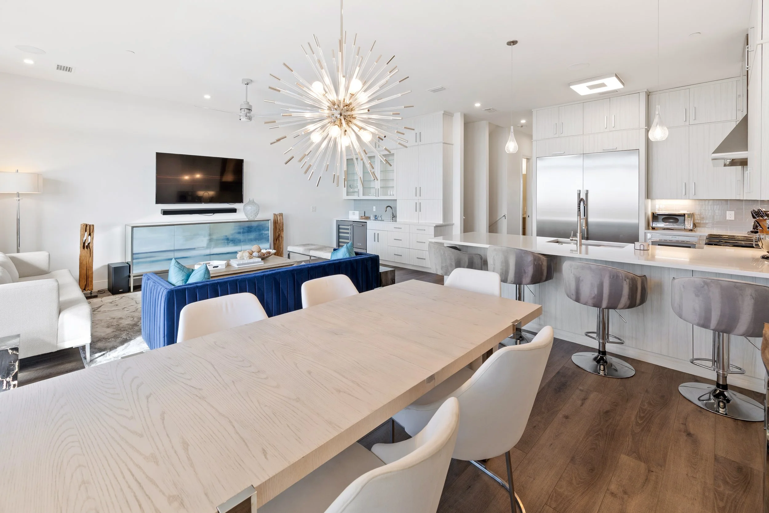 Modern open-concept living room and kitchen with white walls, hardwood floors, and contemporary furniture. Features include a large dining table with white chairs, a blue velvet sofa, a flat-screen TV on a white entertainment unit, and a kitchen with