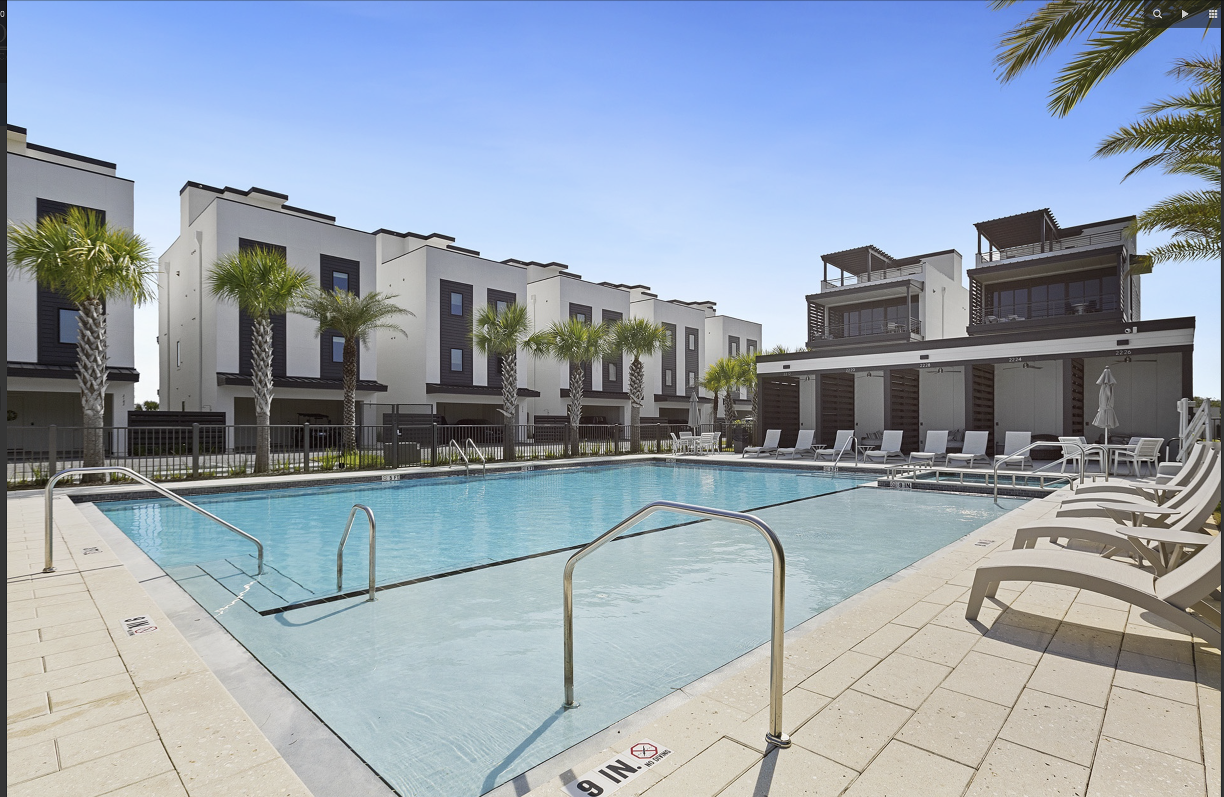 Modern luxury neighborhood with a swimming pool, surrounded by lounge chairs and palm trees, under a clear blue sky.