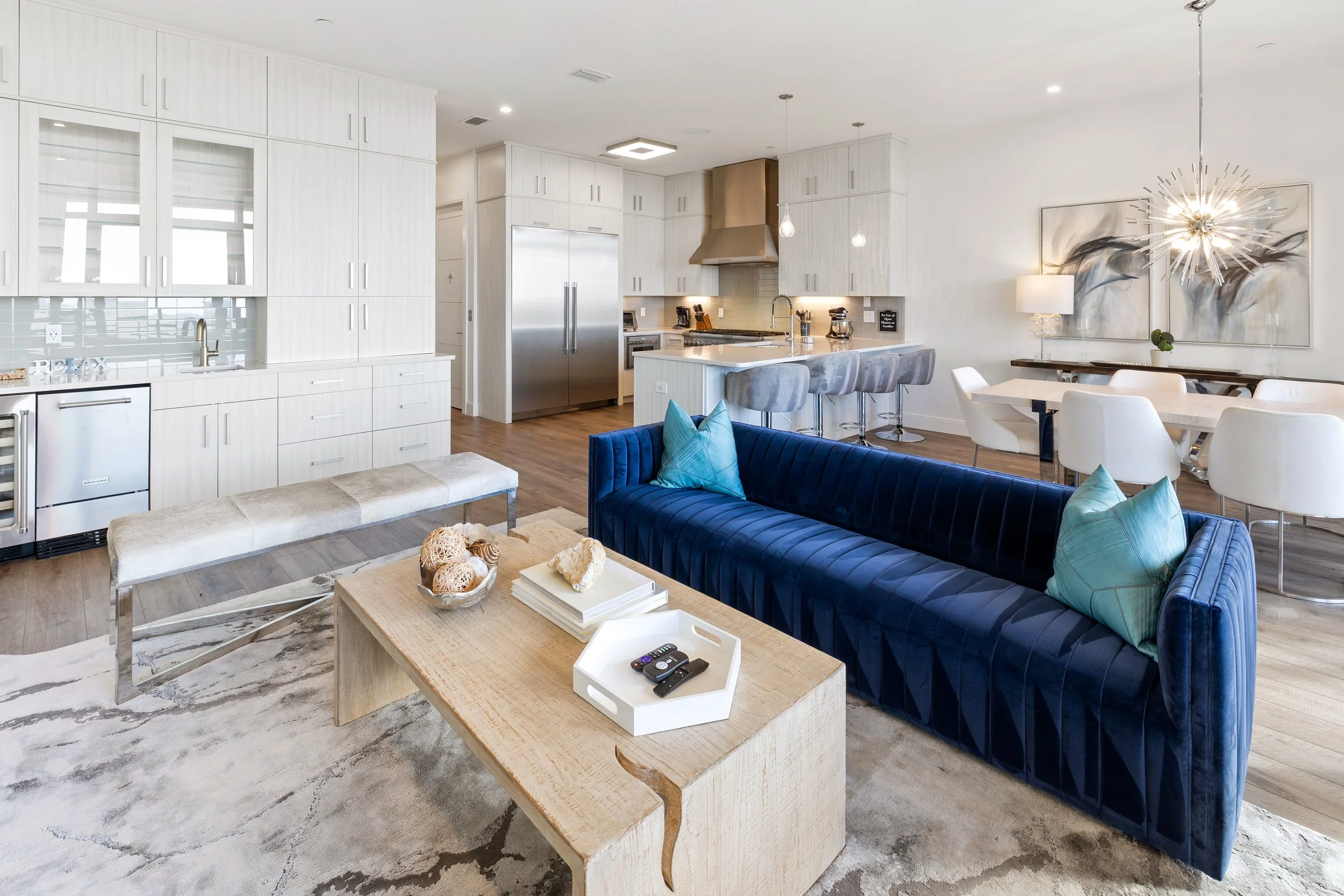 Open-concept living area with a blue velvet sofa, a beige wooden coffee table with remote controls, decorative pillows, a beige bench, and a large painting. The background features a modern kitchen with white cabinetry, stainless steel appliances, an