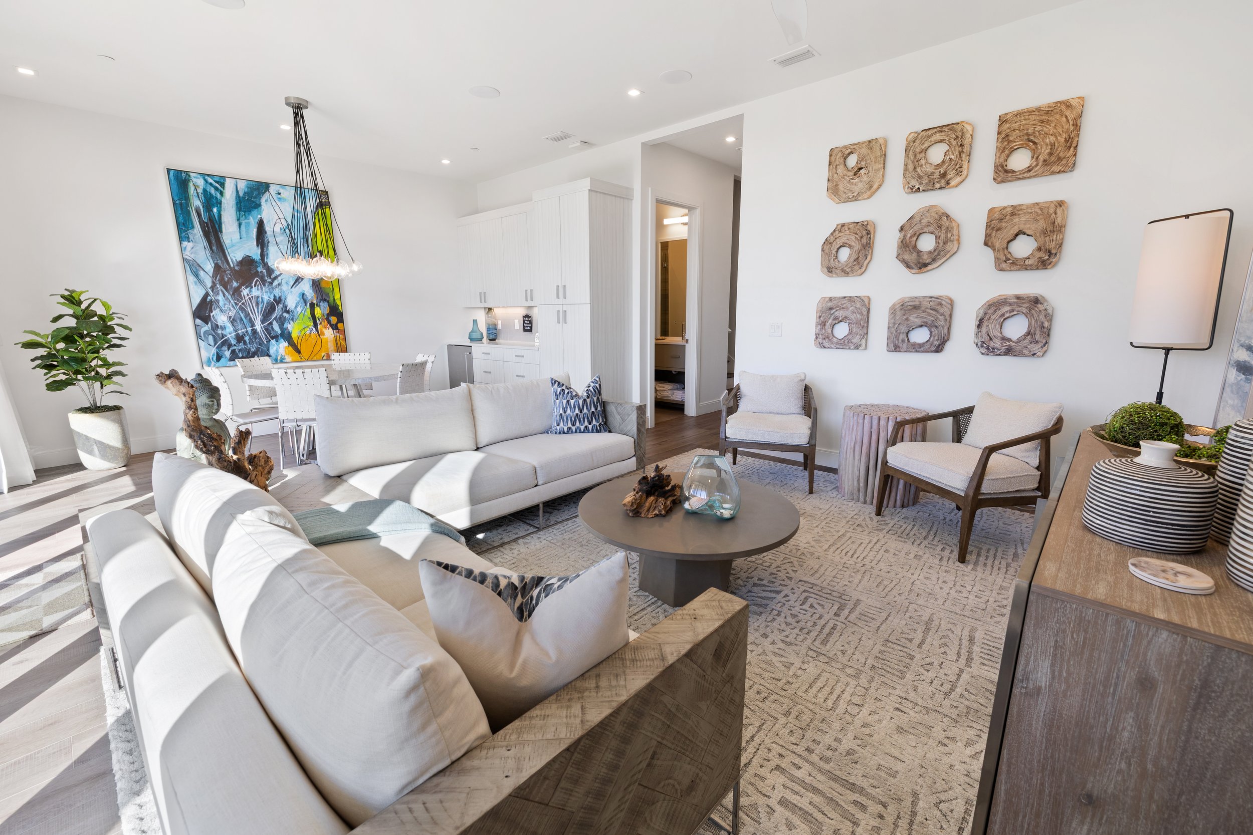 Modern living room with white sofas, wooden chairs, a wooden sideboard, and wall art with wood slices, along with a dining area, decorative vases, and indoor plants.