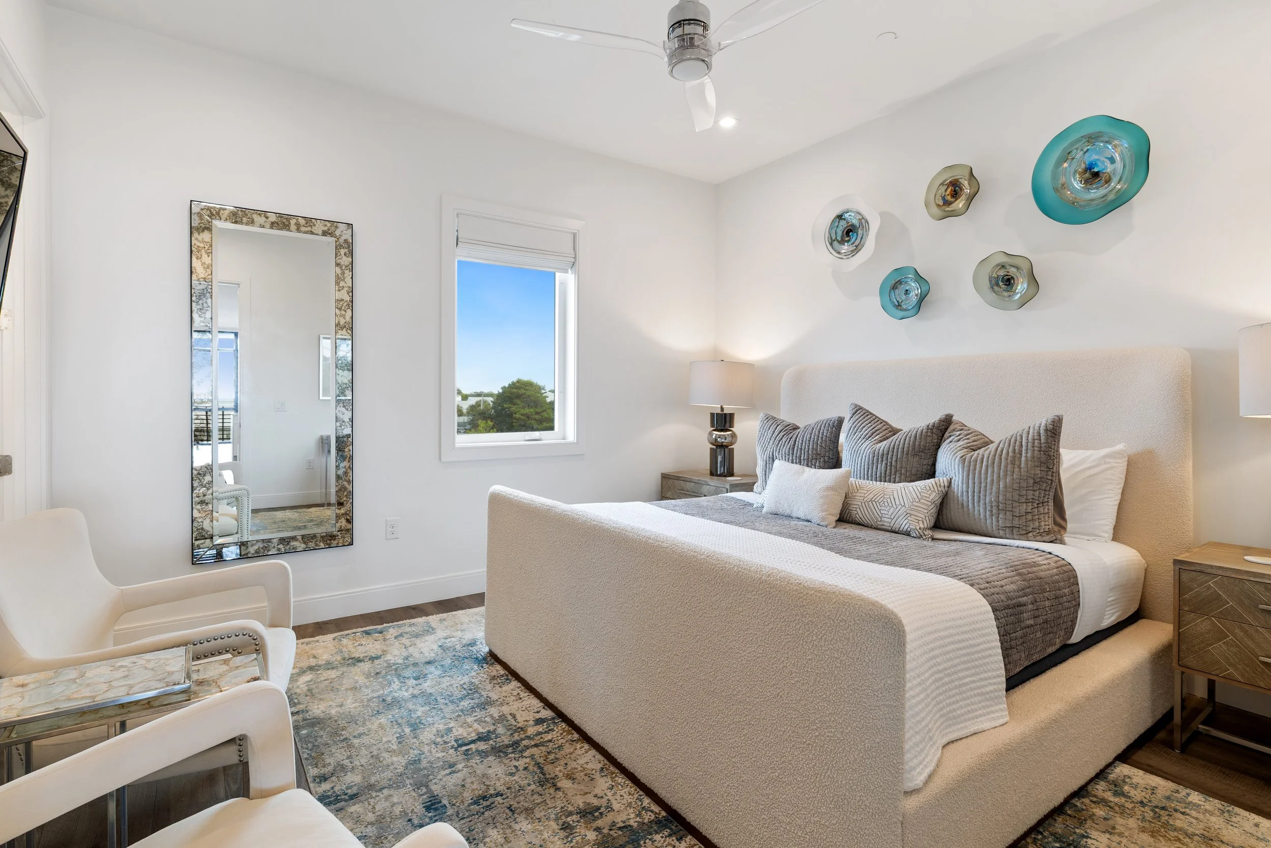 Bright master bedroom decorative gray and white pillows, bedside tables with lamps, a tall mirror, a window with a beautiful view of the swimming pool.