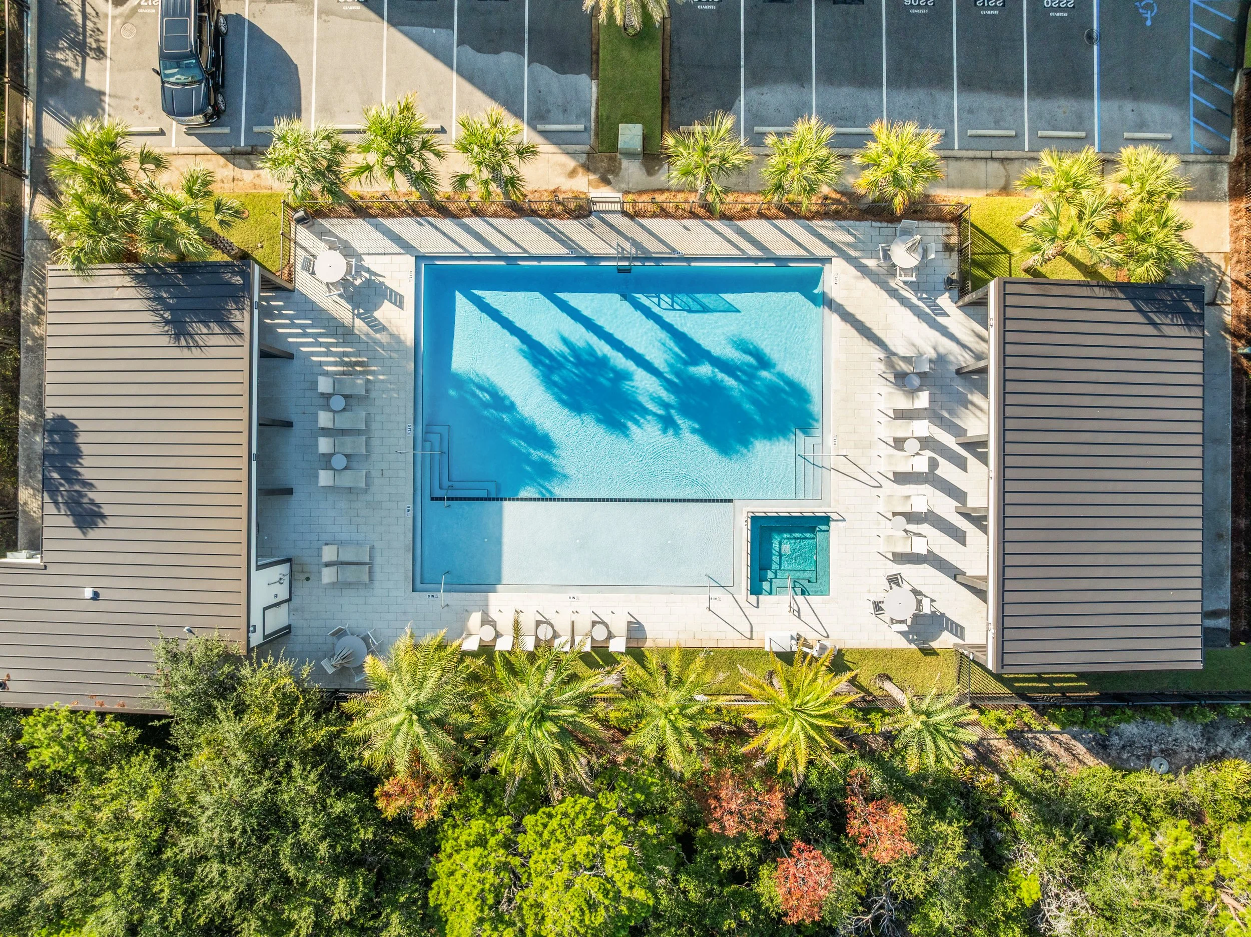 Aerial view of a swimming pool area with lounge chairs, umbrellas, and a hot tub, surrounded by palm trees and parking lot with parked cars.